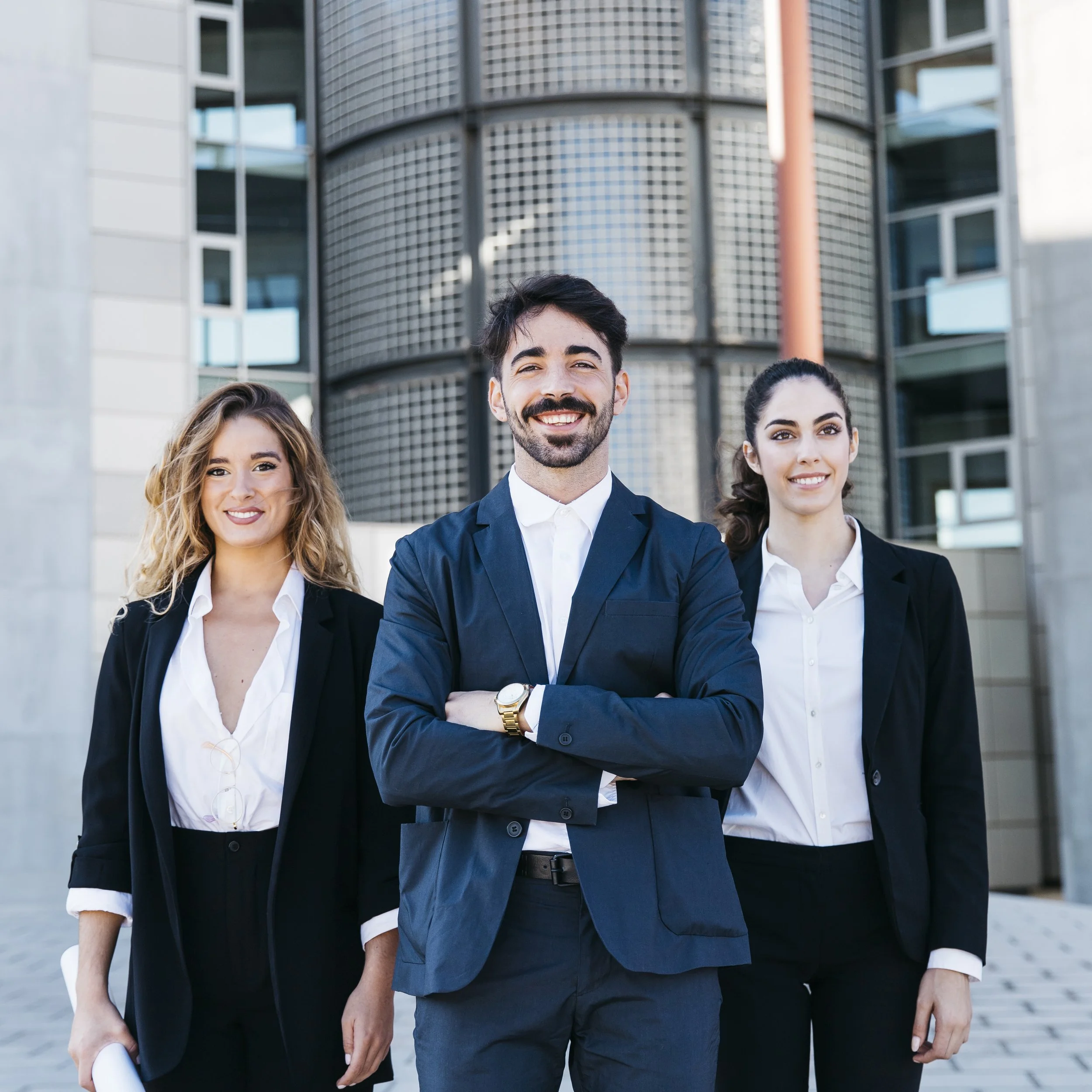 Three business people standing outdoors in front of a modern office building, smiling and looking confident.