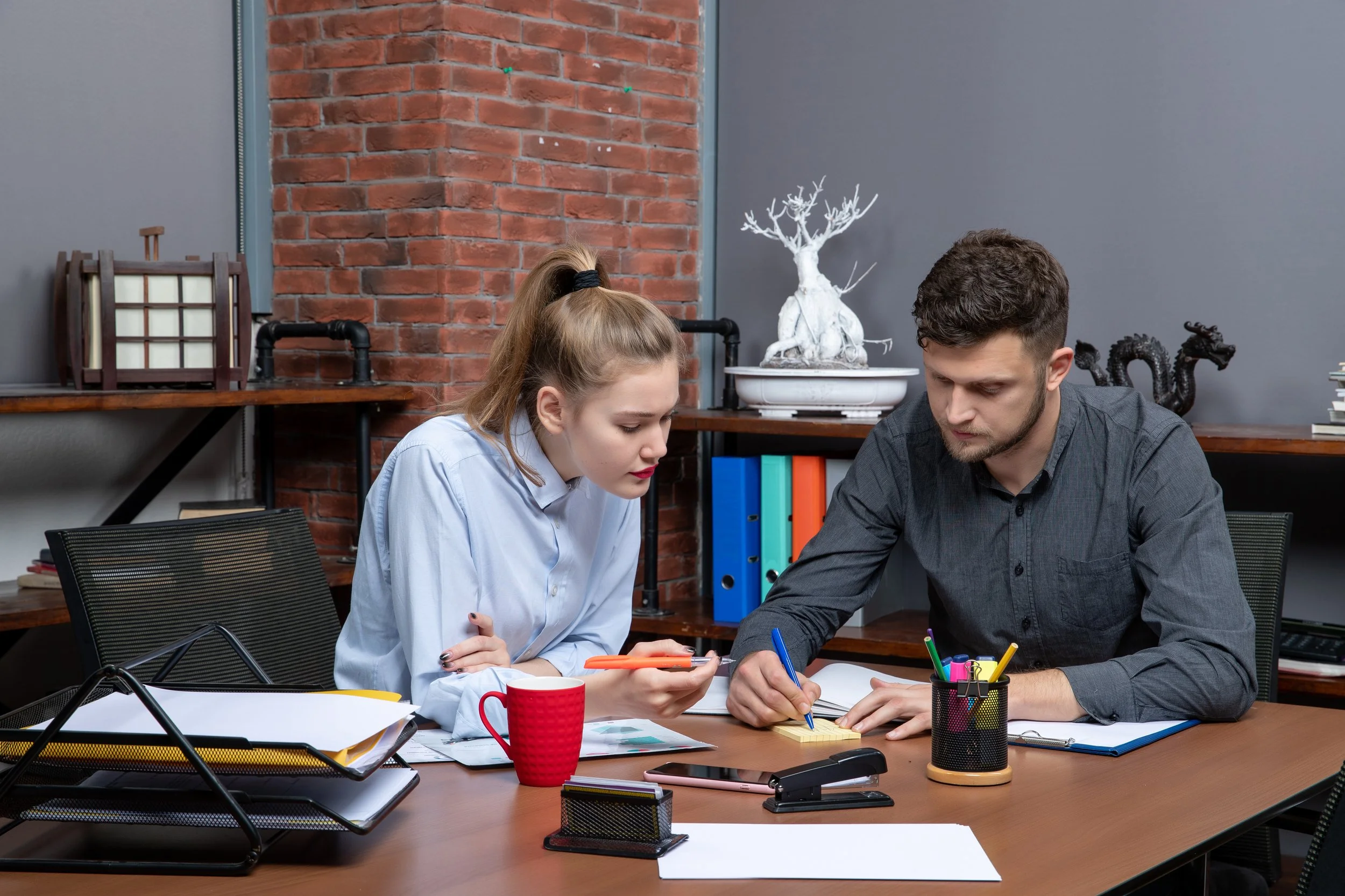 Two people working together at a cluttered office desk, with folders, papers, and writing supplies, in front of a brick wall with decorative objects.