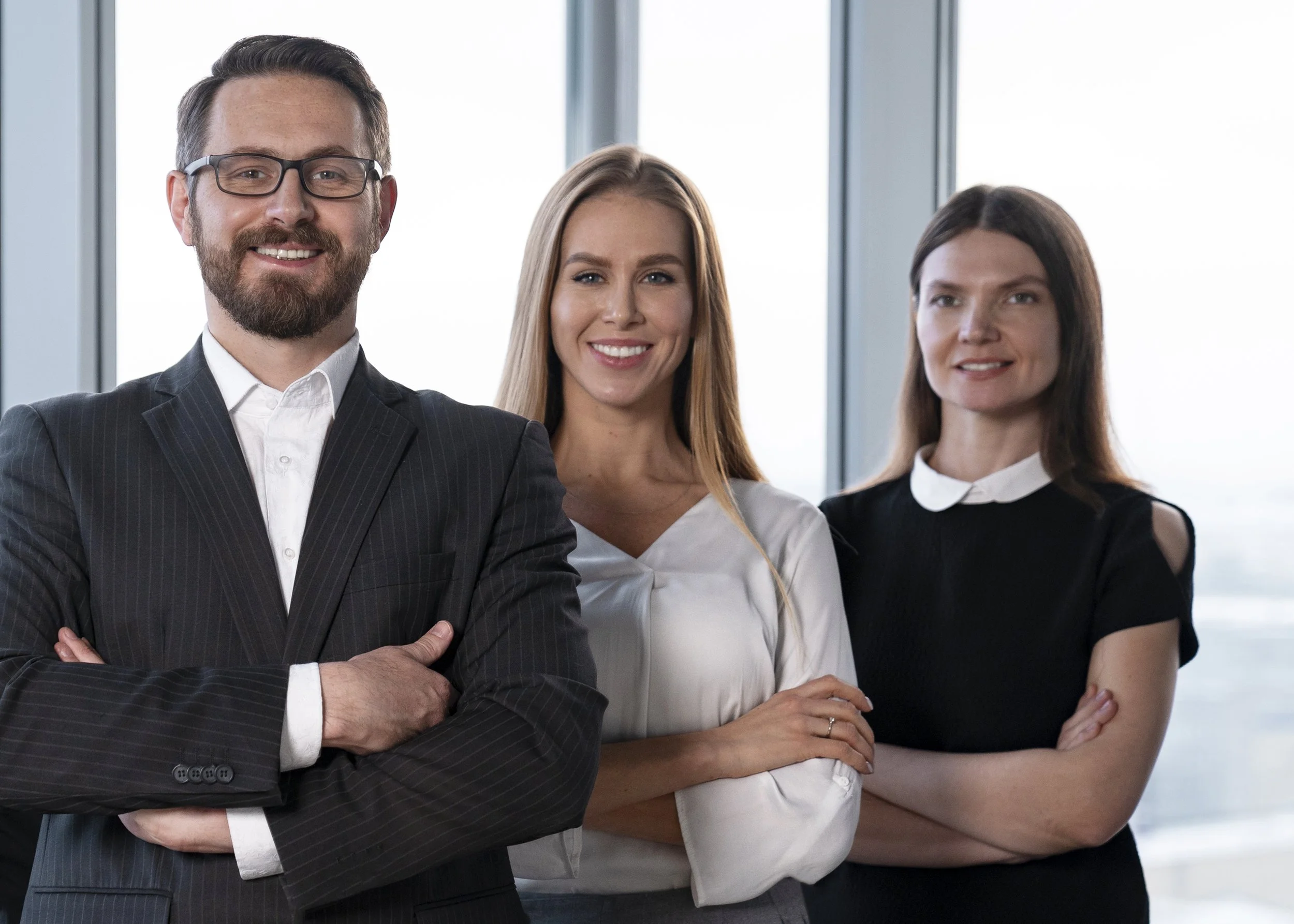 Three professionally dressed people standing with arms crossed inside an office building near large windows, smiling at the camera.