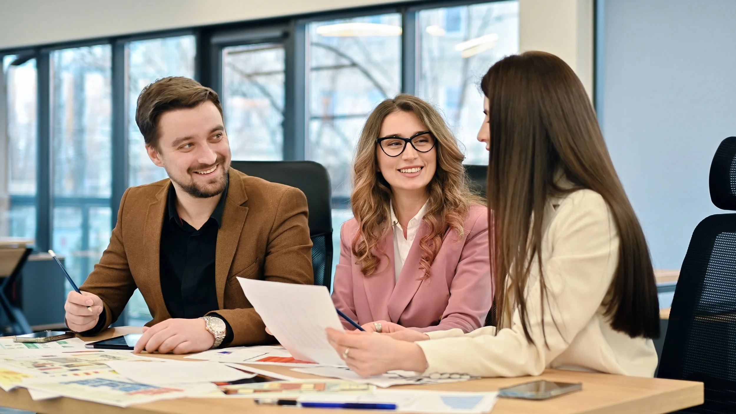 Three business professionals having a meeting in an office, smiling and discussing documents at a desk covered with papers and charts.