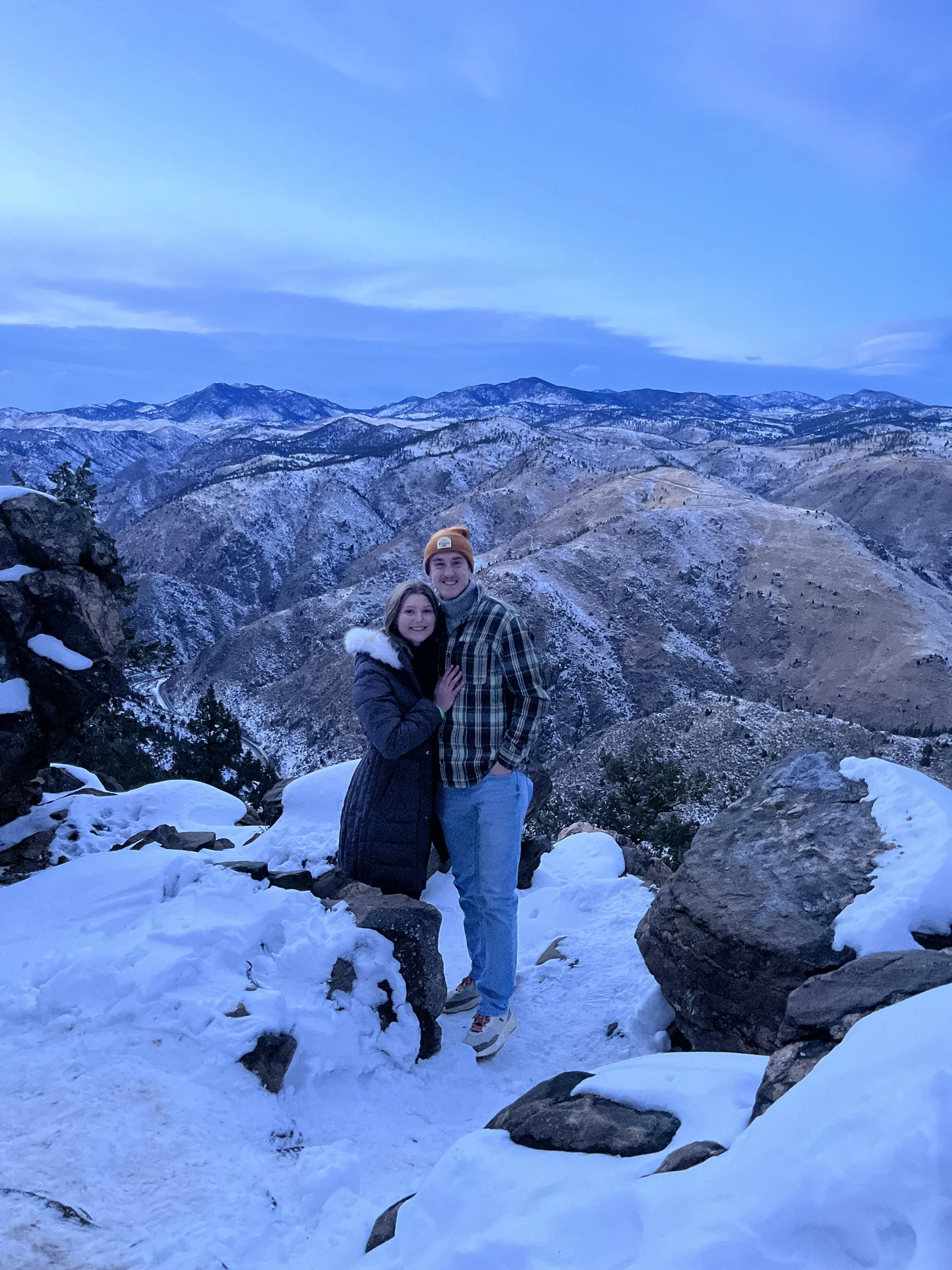 A couple standing on snow-covered rocks in a mountainous landscape with snow on the peaks and a blue sky.