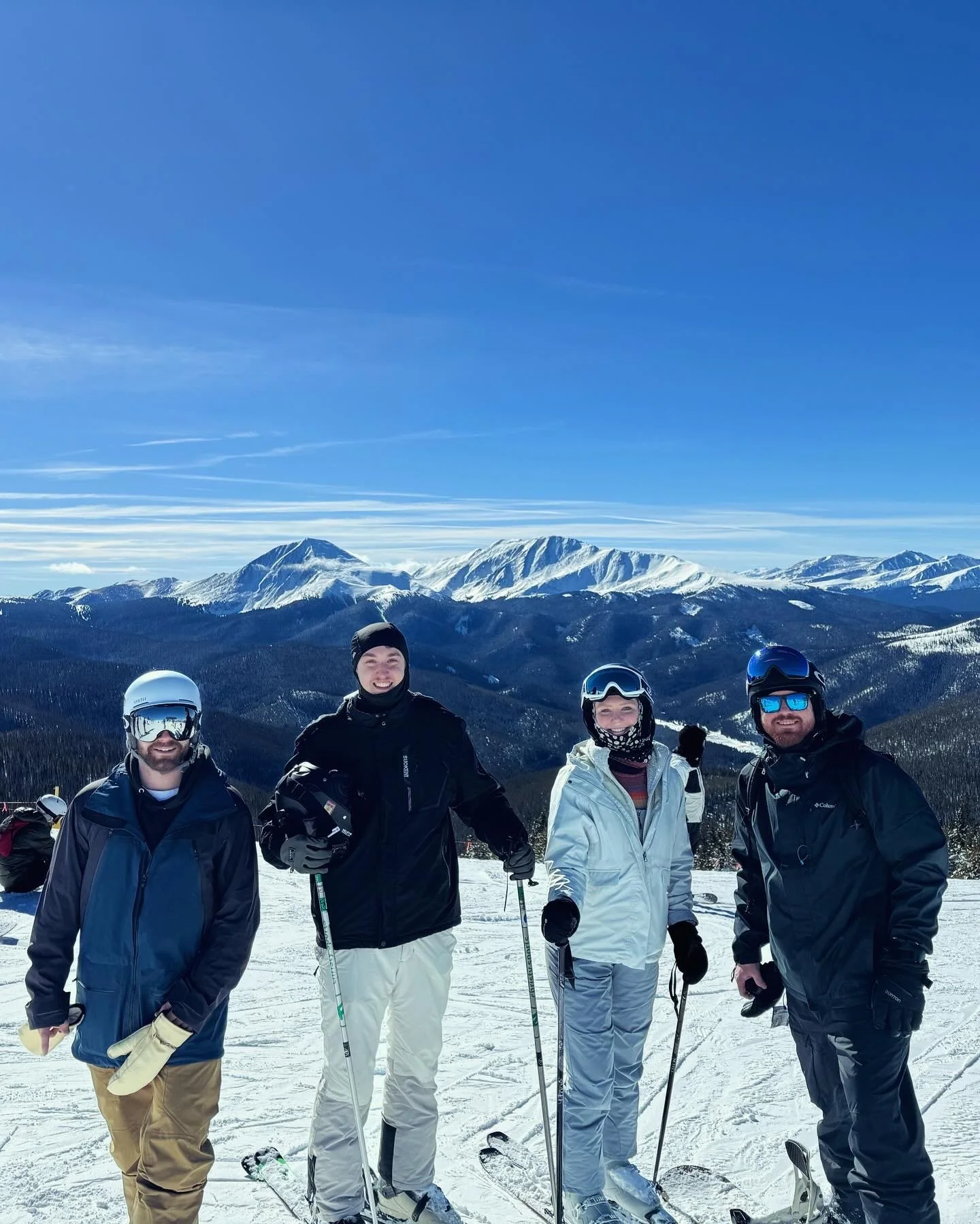 Four people in ski gear standing on snow with mountains in the background.