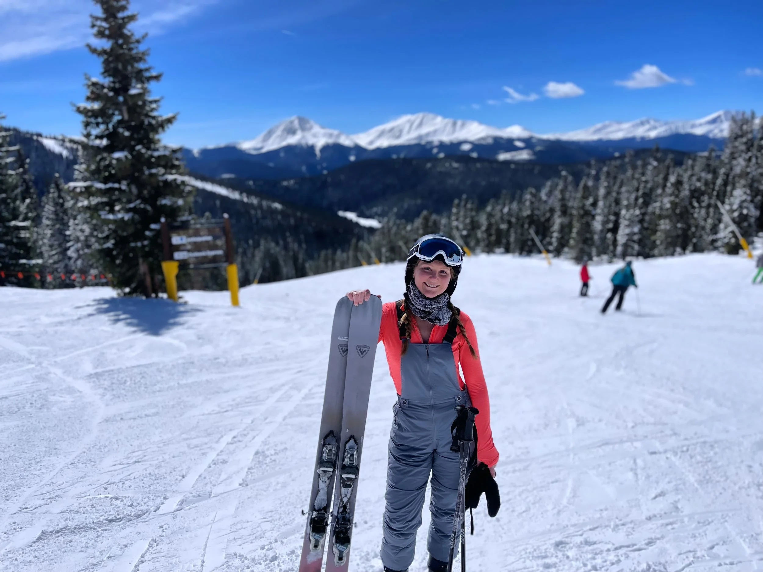 A smiling woman in ski gear standing on a snowy mountain, holding skis and ski poles, with a backdrop of snow-covered trees and mountain peaks under a blue sky.