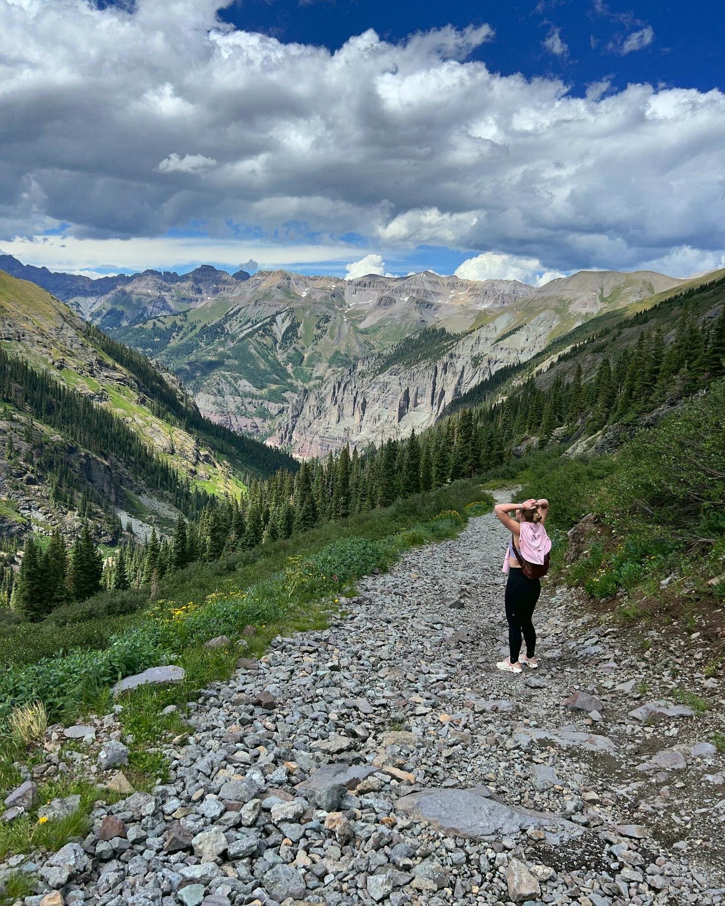 A woman stands on a rocky mountain trail, looking at a scenic view of lush green valleys, dense pine forests, and rugged mountain peaks under a partly cloudy sky.