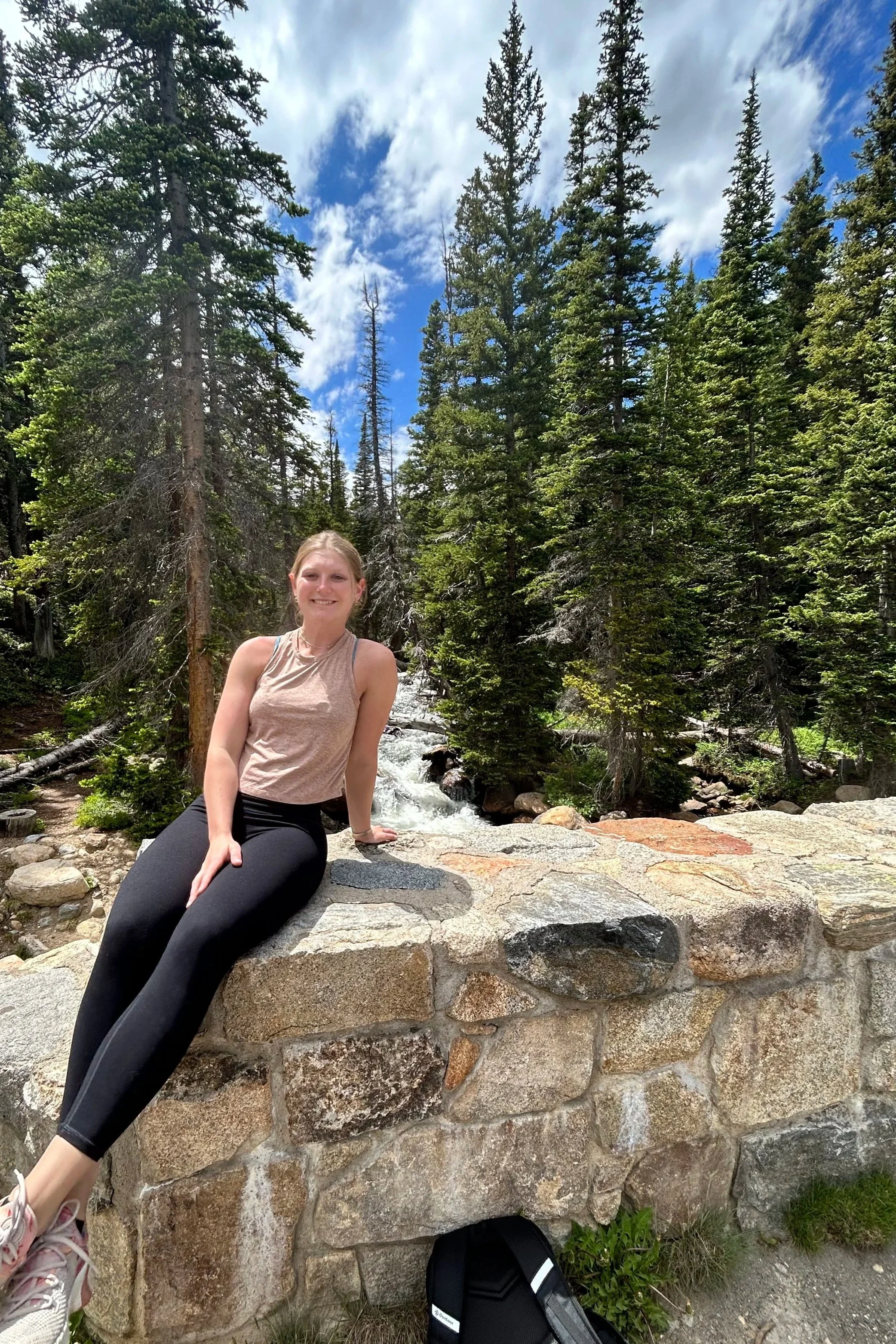 A woman sitting on a stone wall in a forested area with tall pine trees, a small waterfall, and a partly cloudy blue sky in the background.