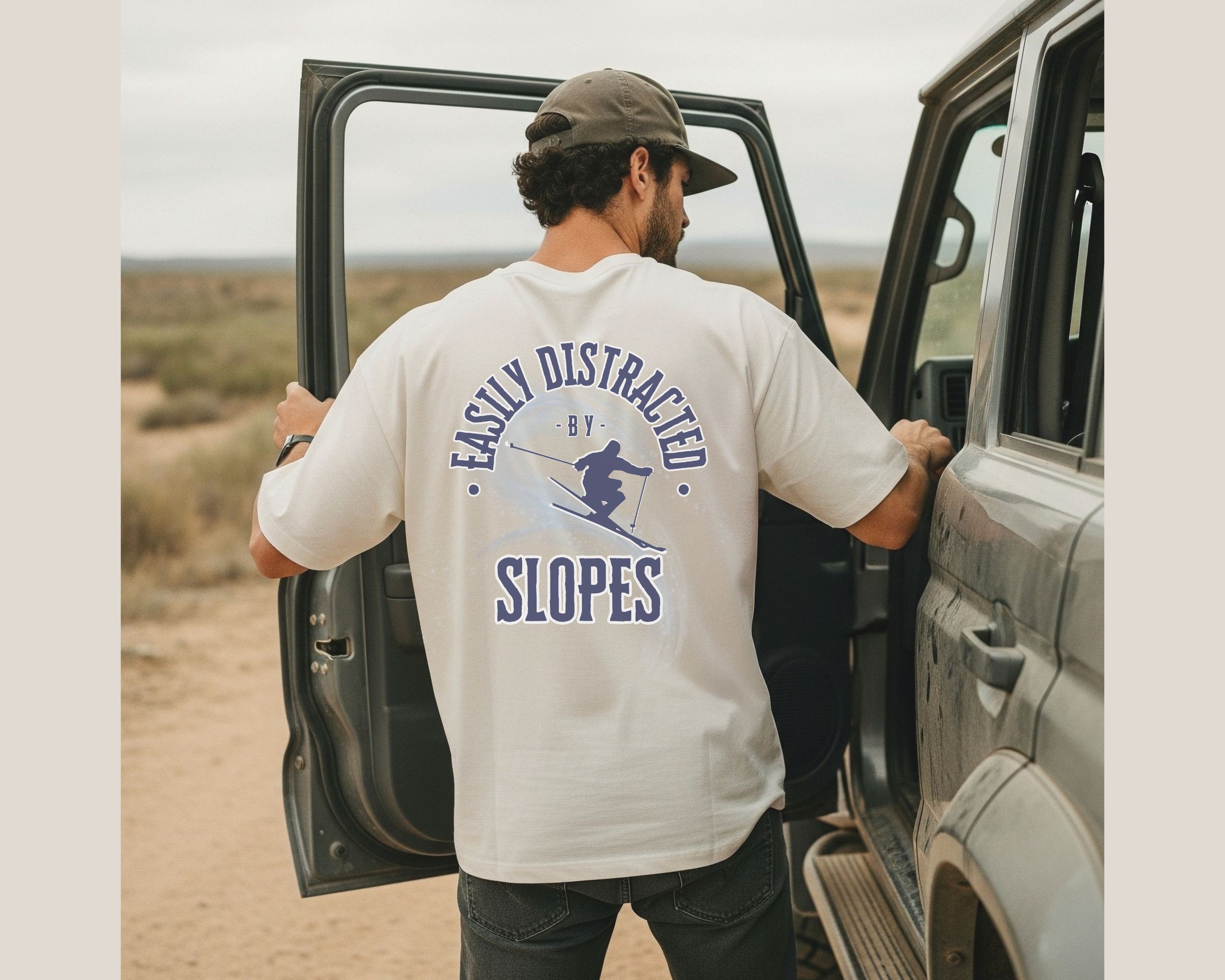 Man loading gear into the back of a gray SUV on a dirt road in a desert landscape.