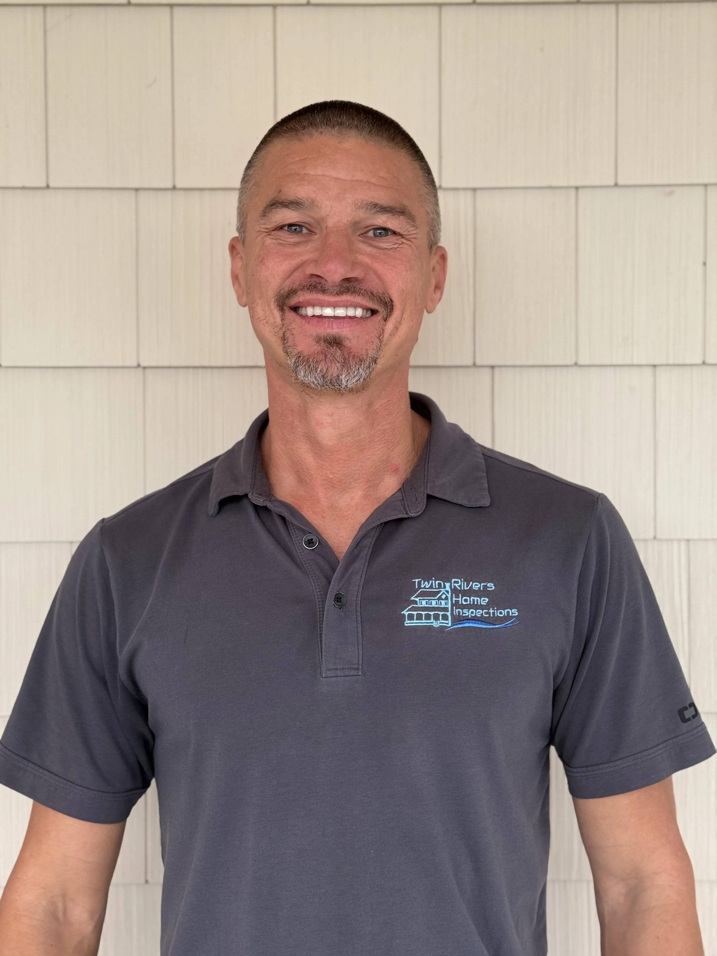 A man smiling, wearing a gray polo shirt with a logo that reads 'Twin Rivers Home Inspections,' standing in front of a white shingled wall.