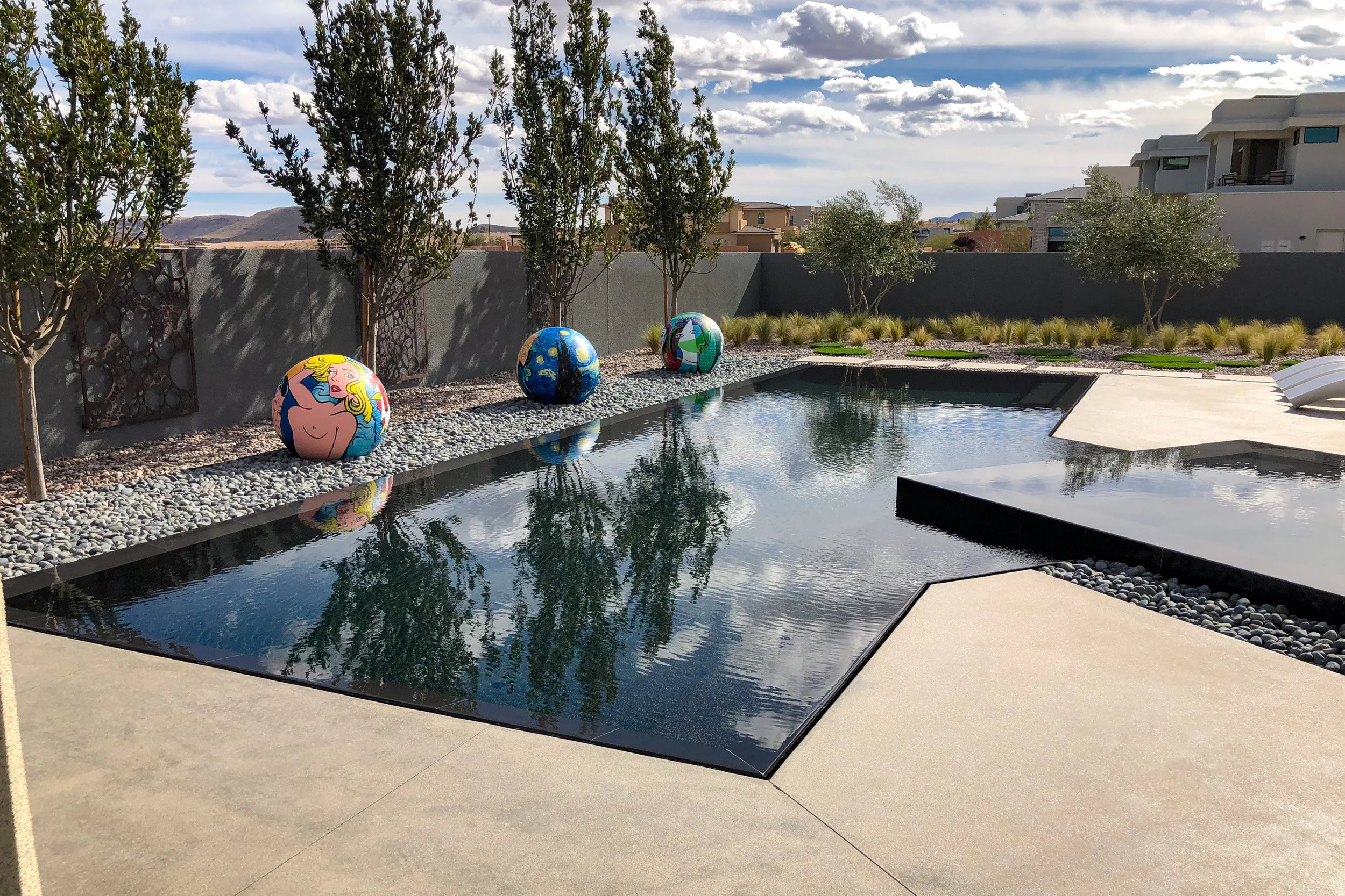 Modern backyard with rectangular swimming pool, decorative art balls, trees, and mountains in the background under a partly cloudy sky.
