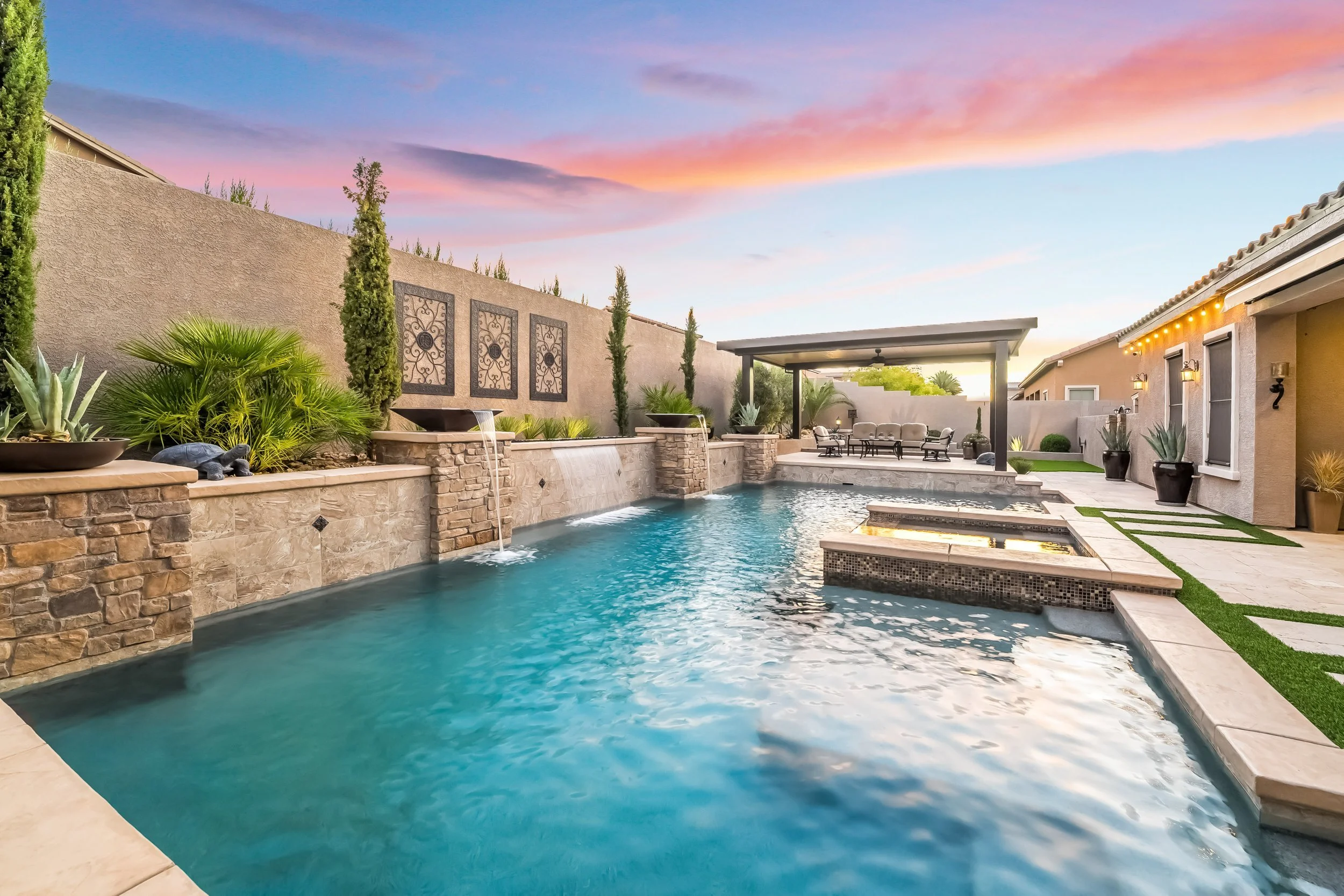 A backyard swimming pool with a waterfall feature, lounge seating area, and decorative plants at sunset.