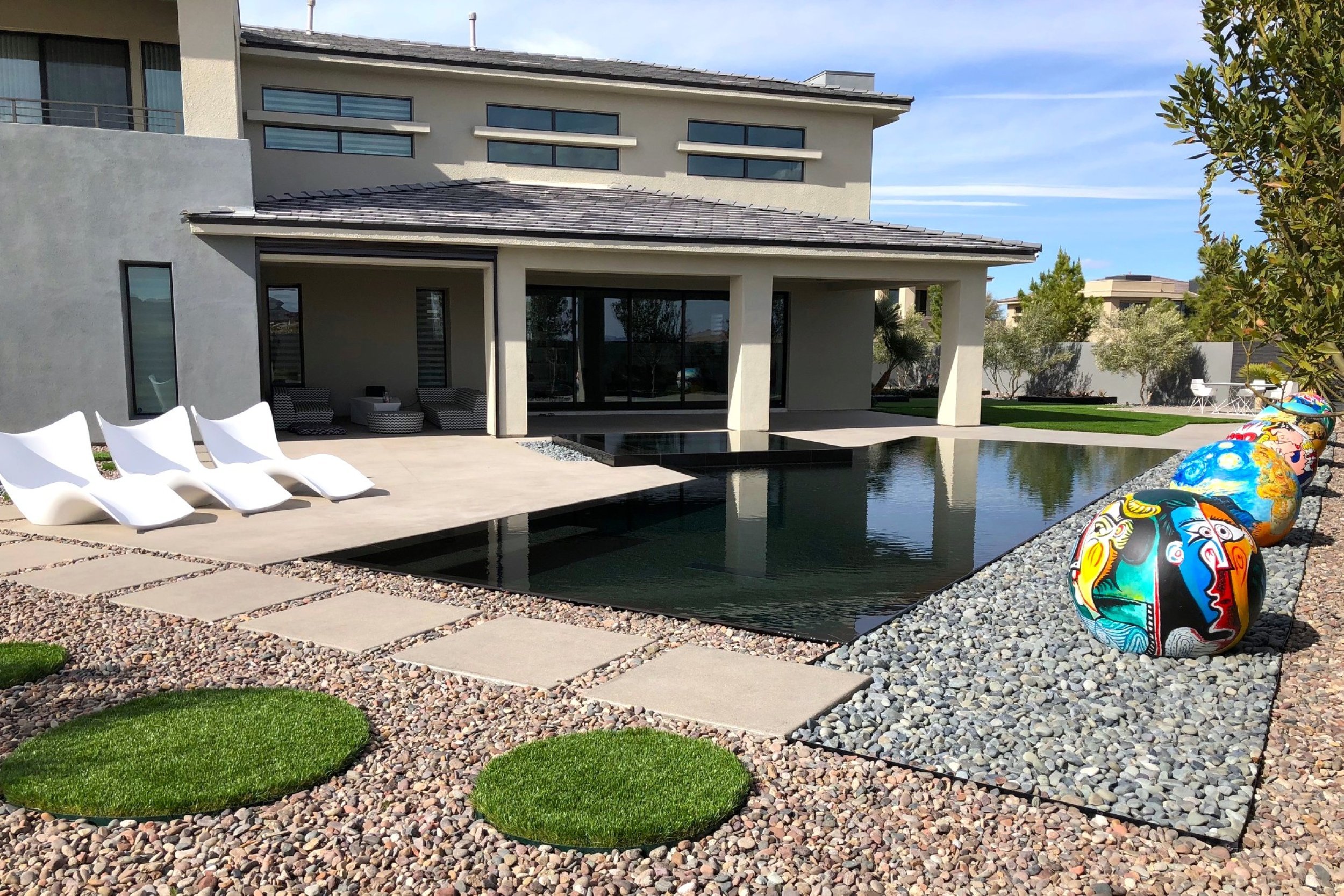 Modern backyard with a rectangular pool, four white lounge chairs, colorful painted decorative stones, and a contemporary house with large windows.