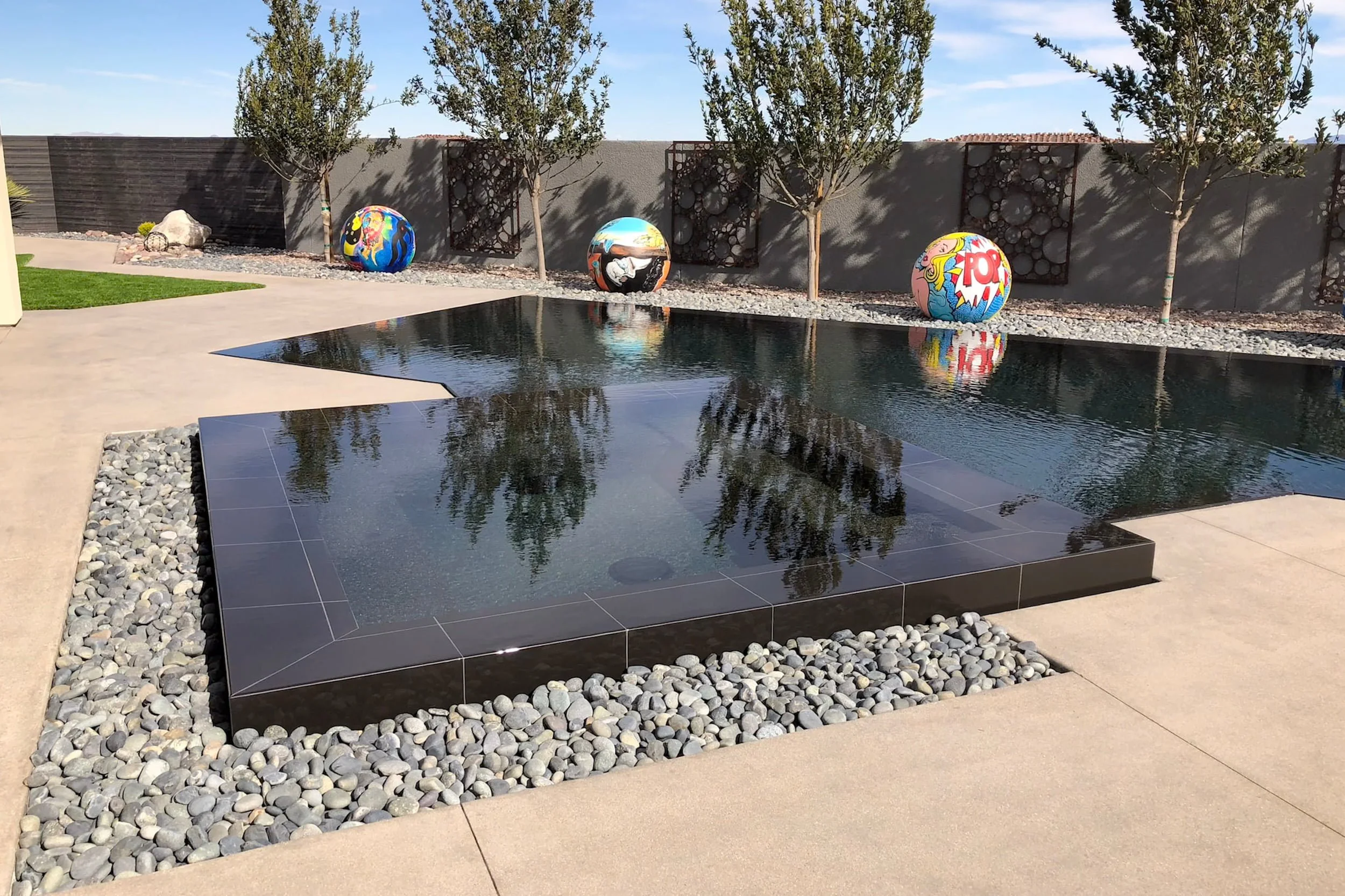 Modern backyard swimming pool with black tile border, surrounded by light concrete patio and gravel. Three colorful decorative spheres with pop art designs and trees along a gray wall in the background.