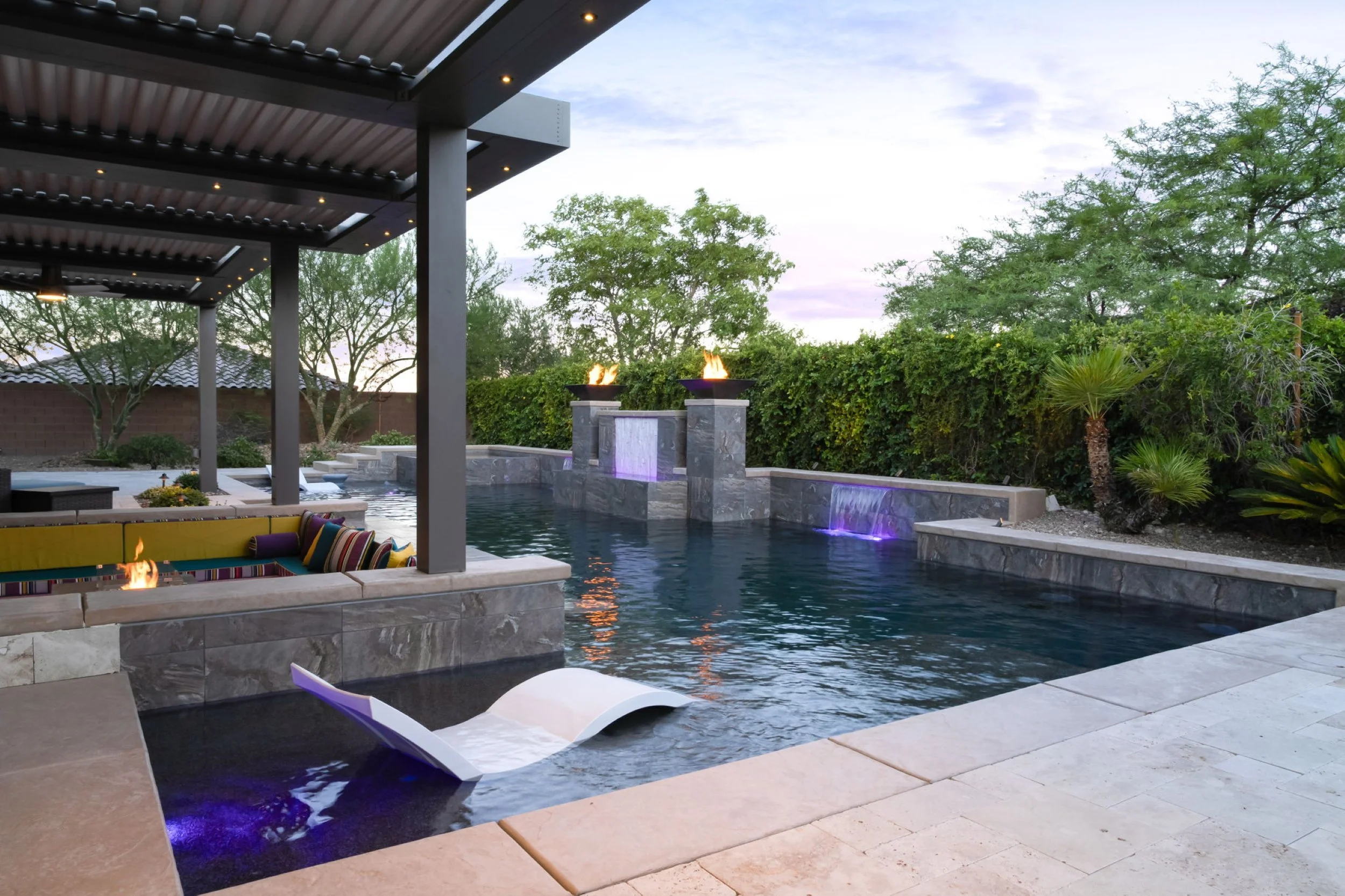 A modern backyard swimming pool with built-in fire pits and water features, surrounded by lush trees and bushes, with a shaded seating area under a structure with string lights, during dusk.