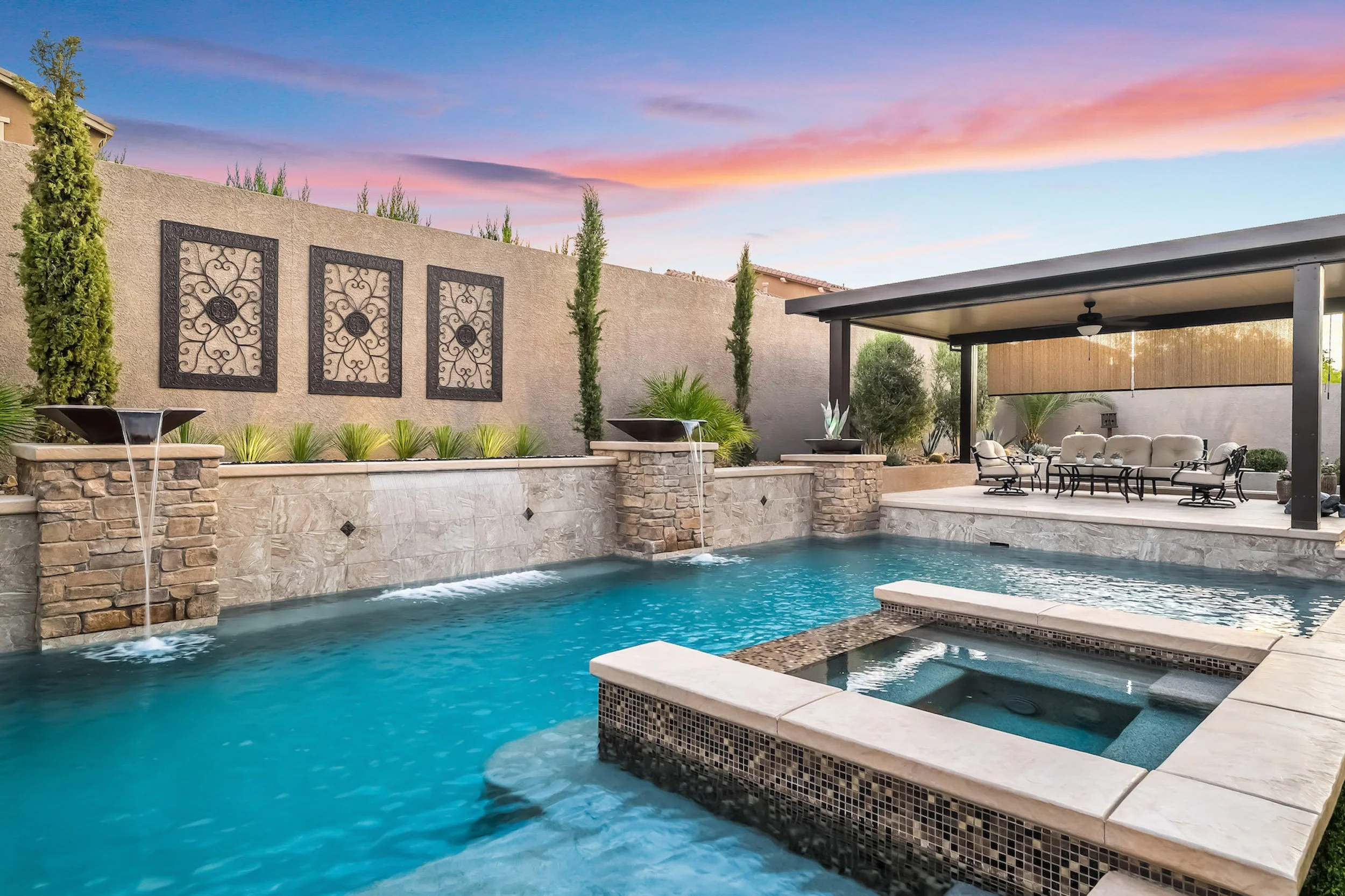 Backyard swimming pool with waterfall features, surrounded by stone and tile, with a covered patio area for seating, with a sunset sky in the background.