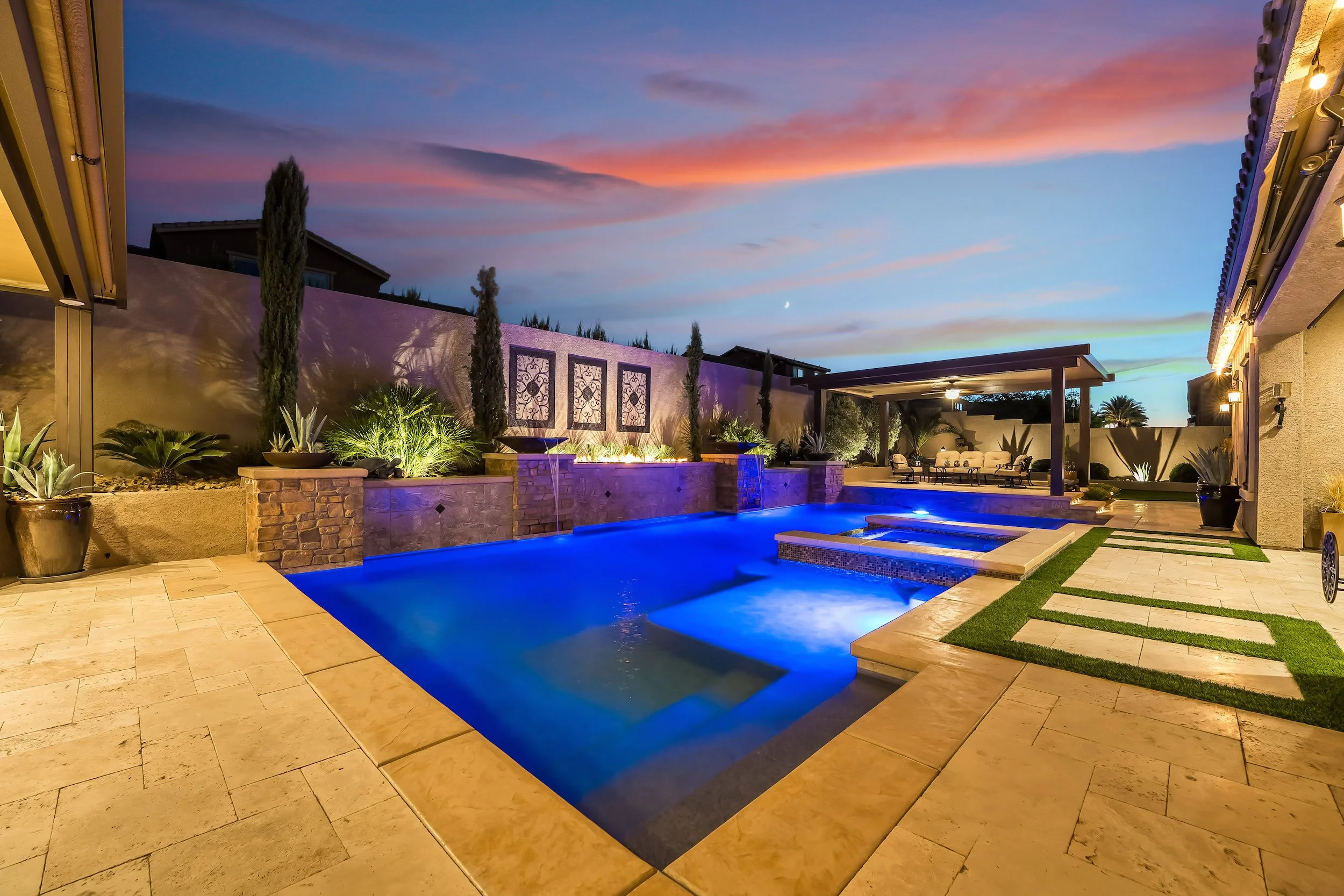 A backyard with a lit swimming pool, patio furniture, and landscaping at dusk.