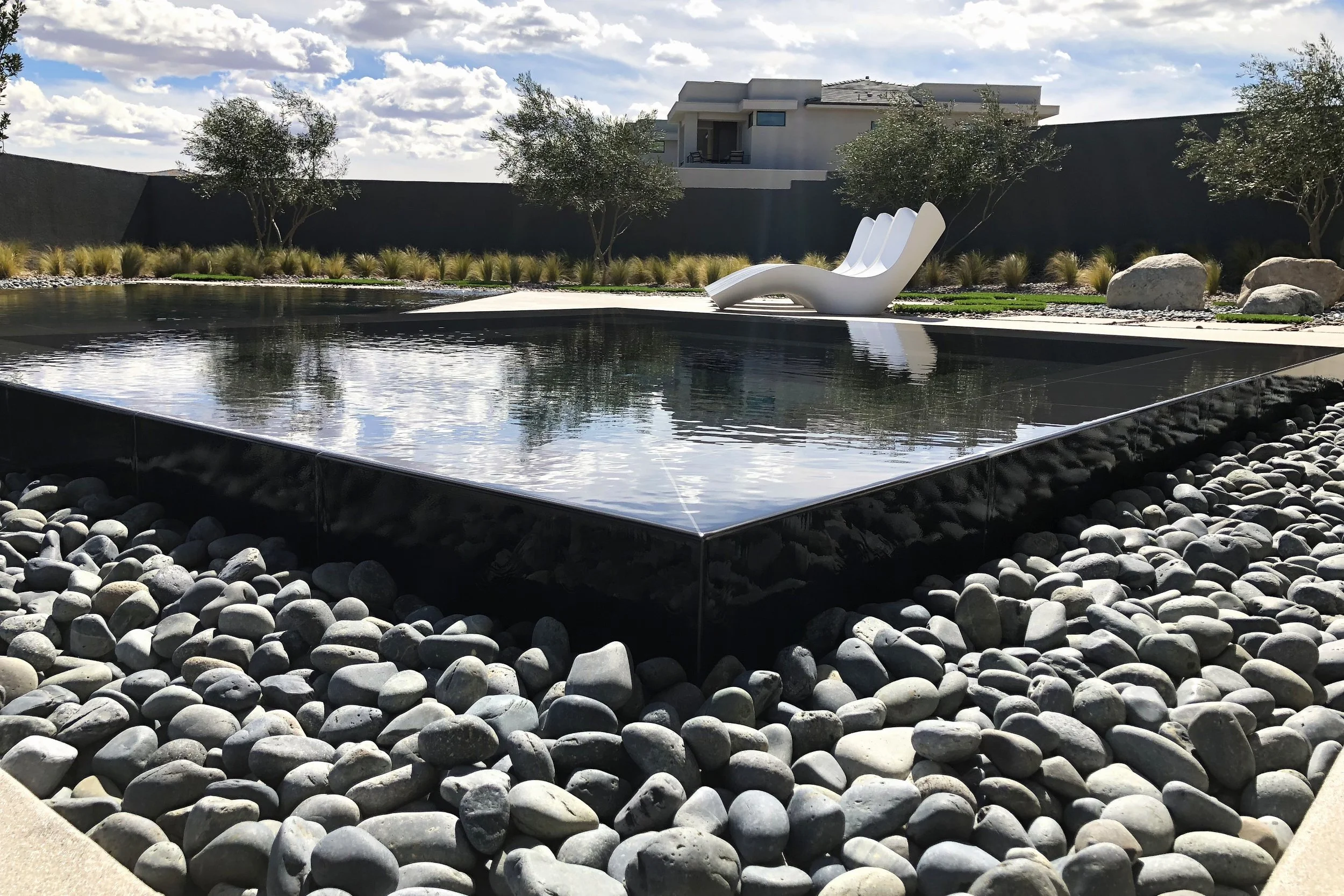 Modern backyard with a rectangular reflecting pool, surrounded by smooth stones, two white lounge chairs, sparse trees, and a contemporary house in the background under a partly cloudy sky.