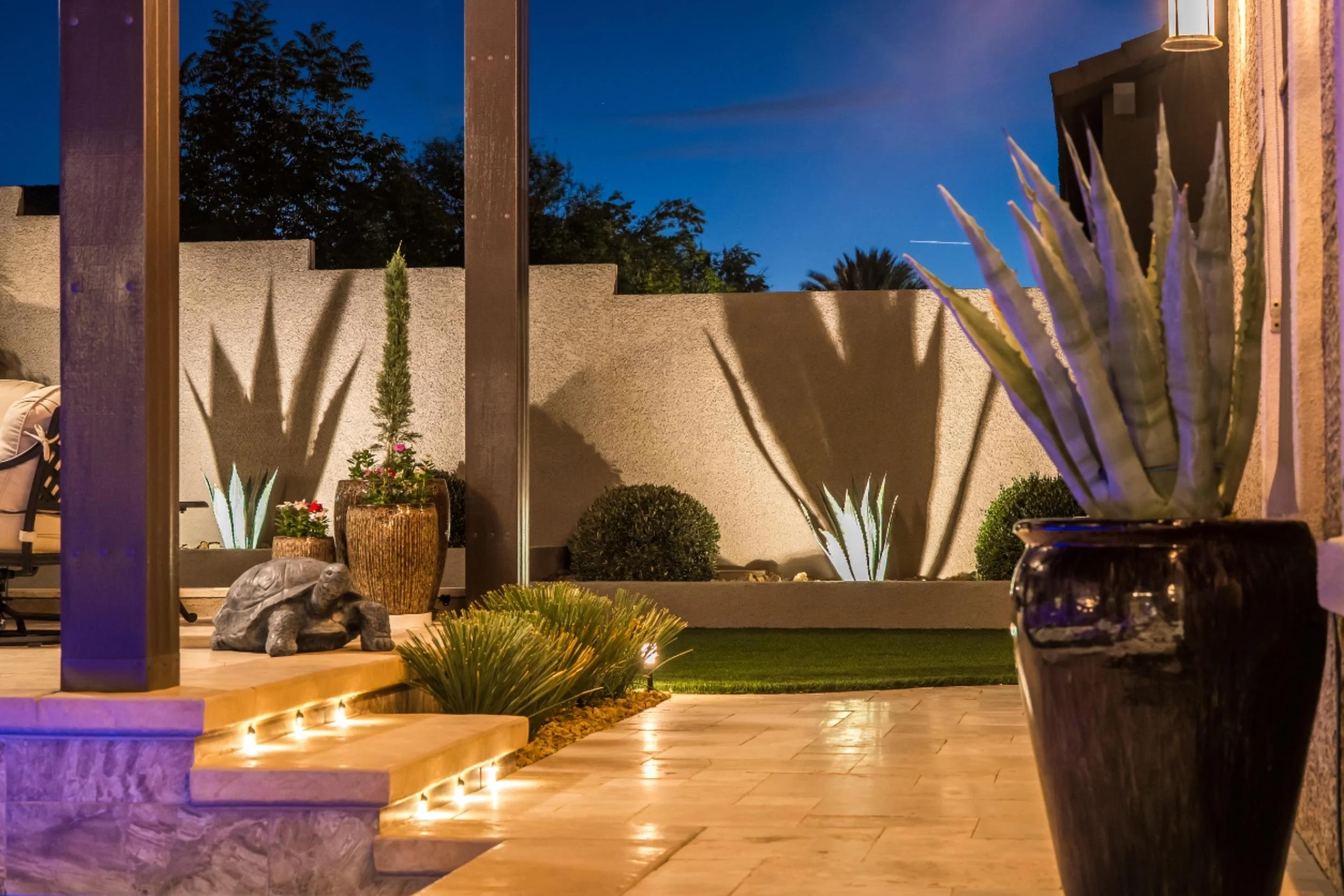 Outdoor patio at dusk with potted succulents, bushes, and decorative lighting, featuring shadows cast by plants on a white wall and a textured raised flower bed.