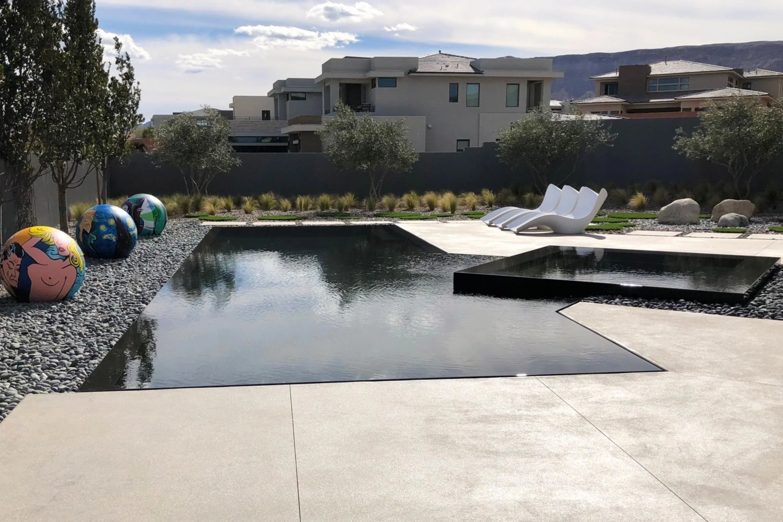Modern backyard with a rectangular reflecting pool, three white lounge chairs, decorative colorful balls, trees, rocks, and mountains in the background.