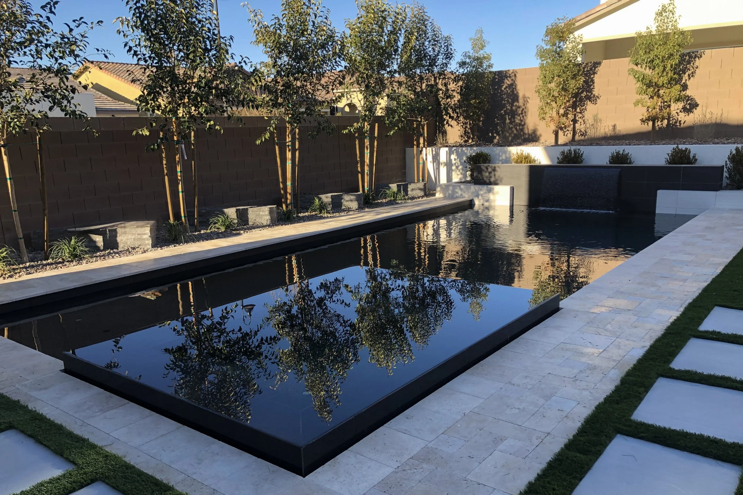 Modern backyard with rectangular black reflecting pool, white stone walkway, featuring evenly spaced square stepping stones, green lawn patches, and a brown privacy wall with trees and small plants.