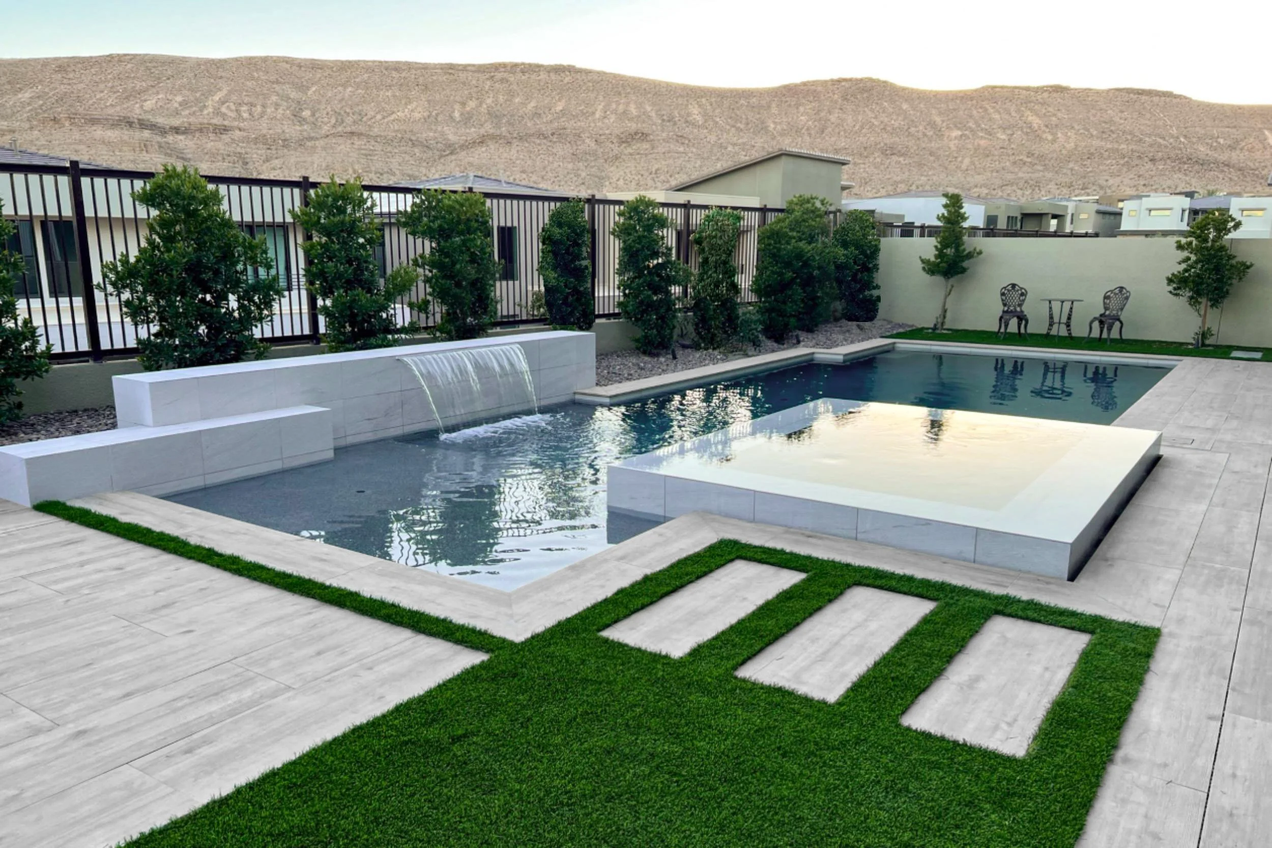 Modern backyard with a swimming pool, water feature, and outdoor furniture, surrounded by a fence and hills in the background.