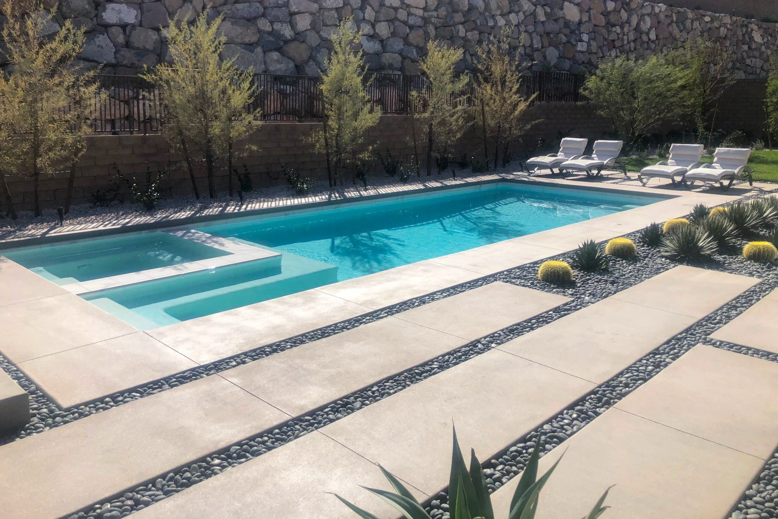 A backyard swimming pool with a built-in hot tub, surrounded by a concrete deck with decorative rocks. There are four lounge chairs along the side, some trees and a stone wall in the background.