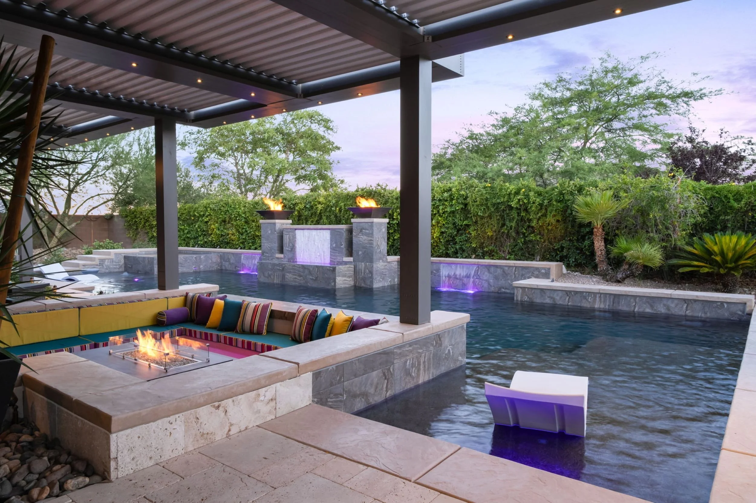 Luxury backyard pool area with a fire pit, lounge chairs, a water feature, and greenery including trees and bushes, during dusk.