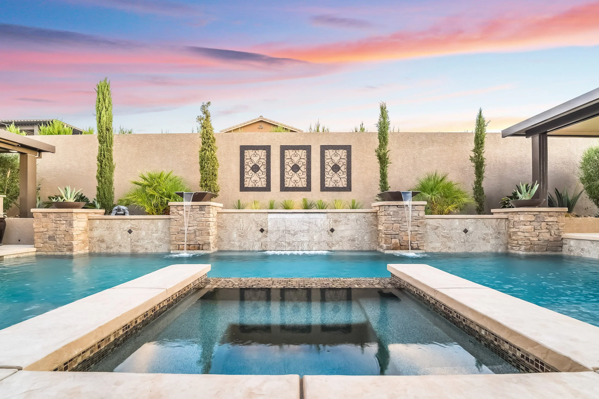 A backyard swimming pool with a hot tub, bordered by stone and concrete, with water features and landscaped plants, a privacy wall with decorative panels, and a sunset sky in the background.