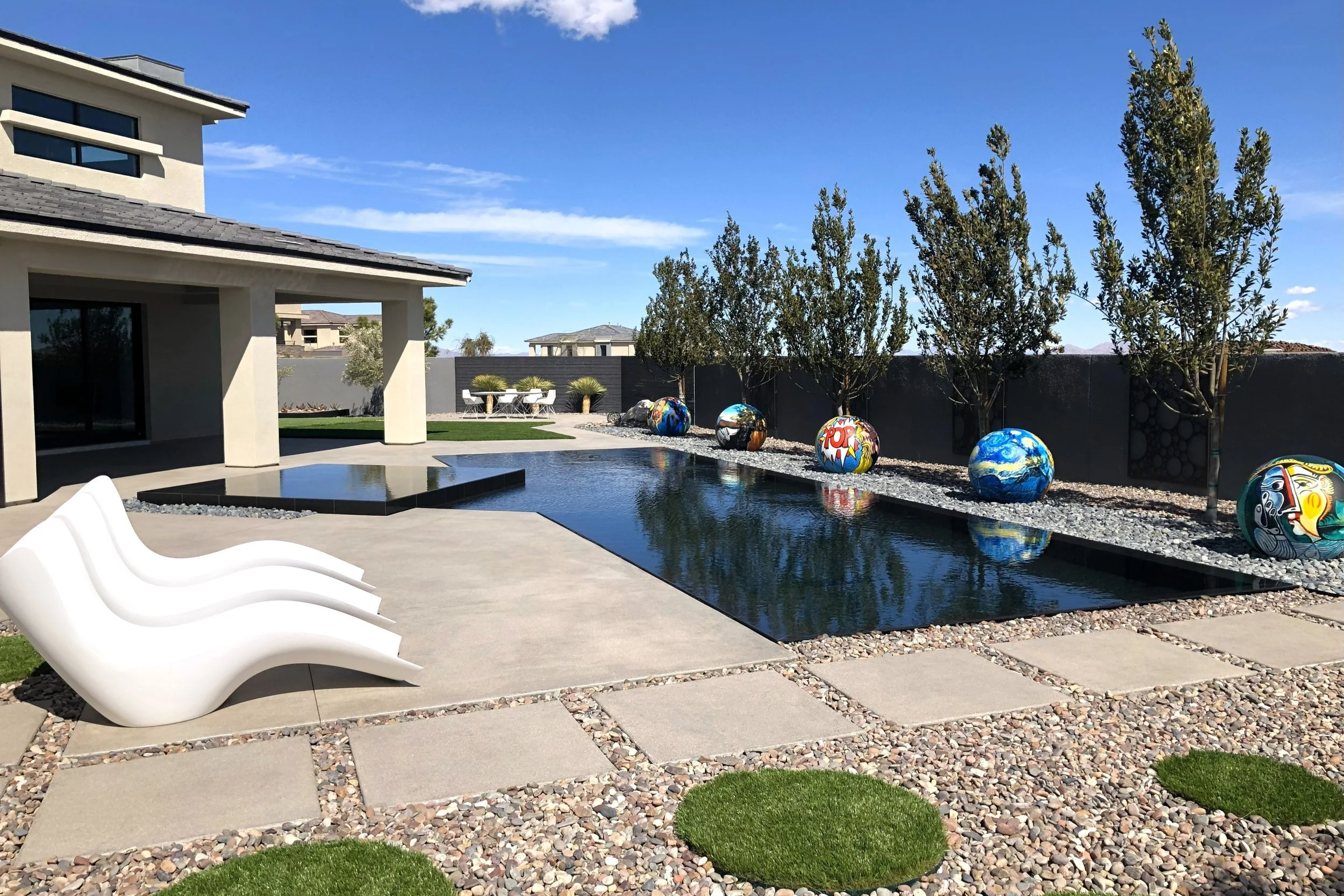 Modern backyard with a rectangular pool, white lounge chairs, a stone patio, and colorful spherical sculptures along a landscaped area with trees.