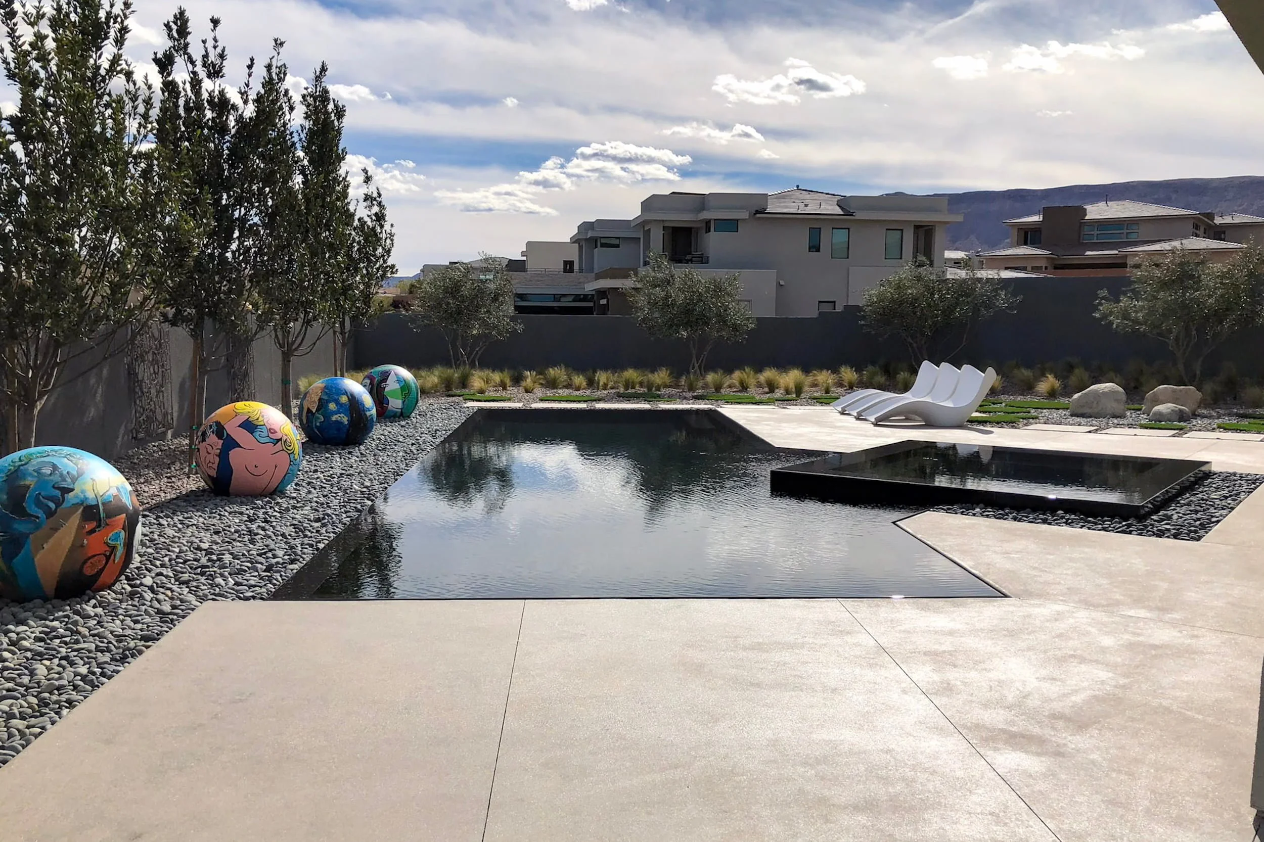Modern backyard with a rectangular swimming pool, decorative painted balls on gravel, white lounge chairs, trees, and contemporary houses in the background under a partly cloudy sky.