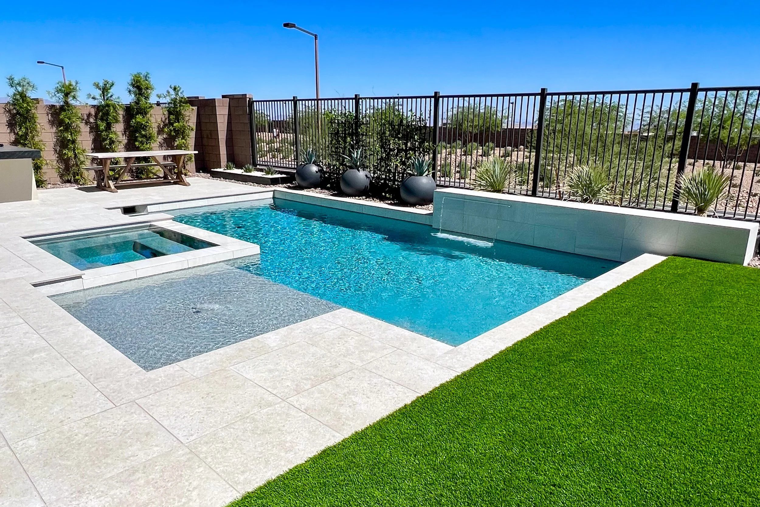 A backyard swimming pool with a small attached spa, surrounded by a tiled deck and green artificial grass, enclosed by a black metal fence, with potted plants and trees in the background under a clear blue sky.