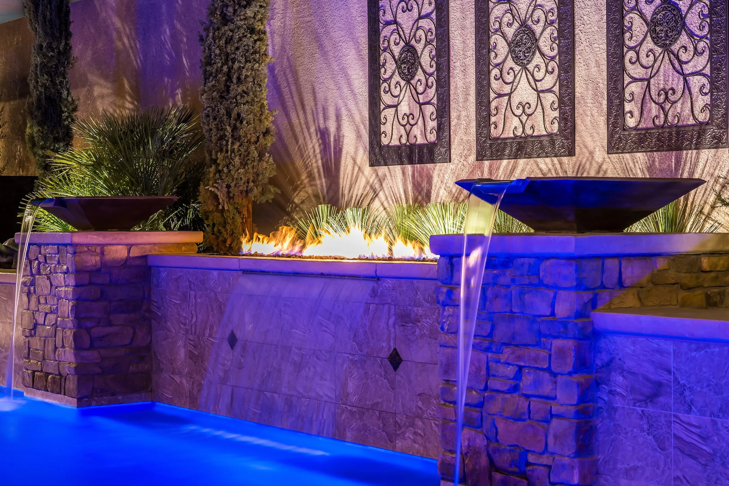 A backyard fire feature with a built-in gas fire pit, surrounded by landscaping with trees, bushes, and decorative wrought iron wall panels, illuminated with purple and blue lighting.
