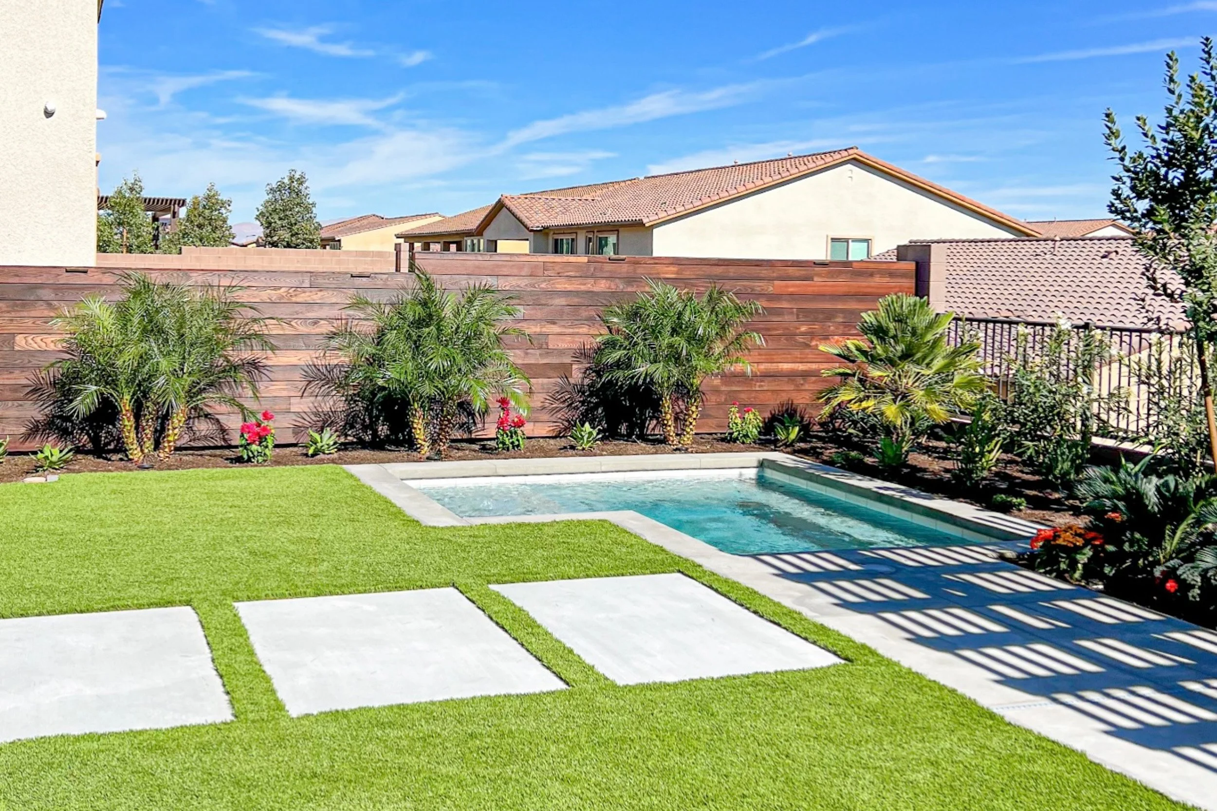 Backyard with a small swimming pool, manicured lawn, tropical plants, a wooden fence, and neighboring houses under a blue sky.