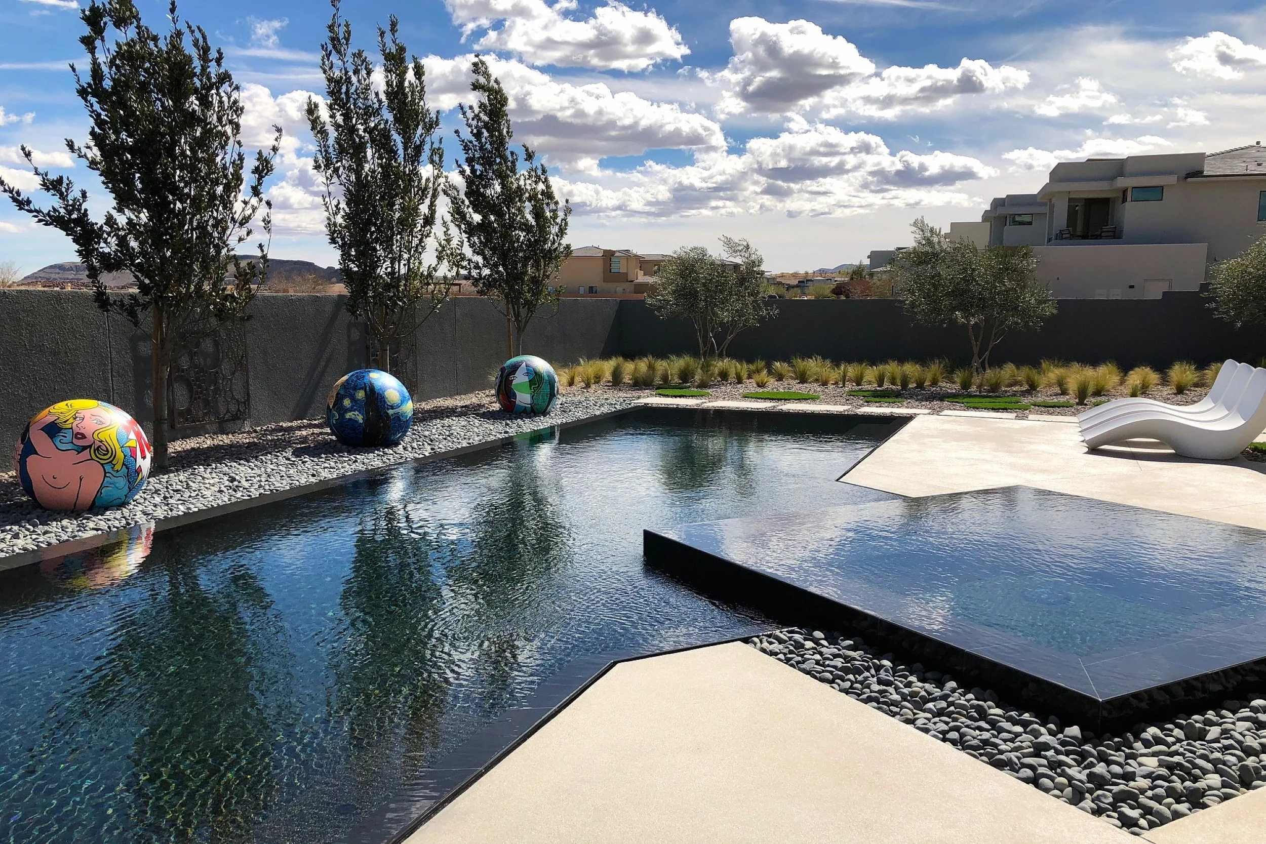 Modern backyard with a sleek pool, decorative spherical art pieces, lounge chairs, trees, and landscaped garden with grass and plants, under a partly cloudy sky.