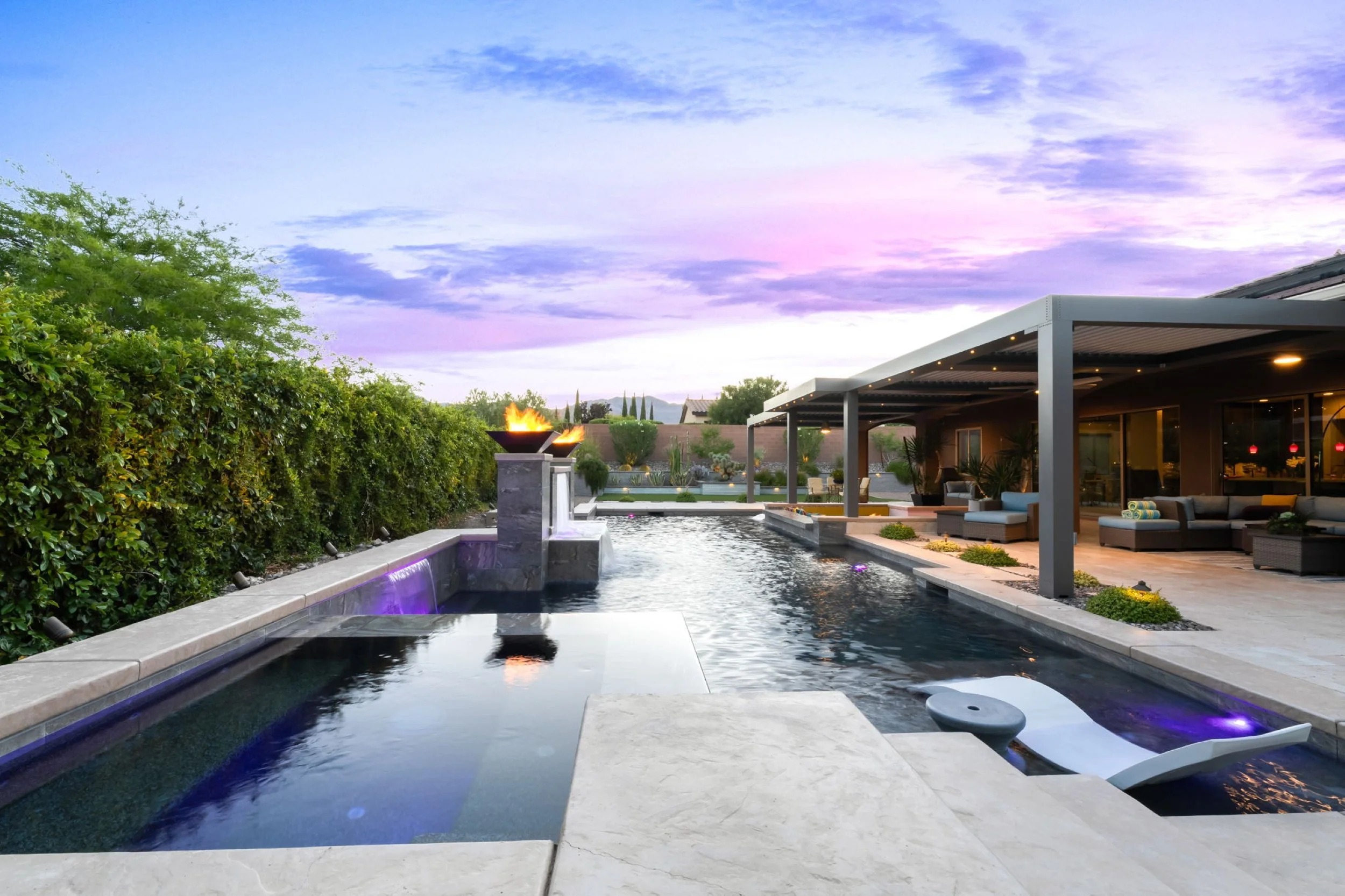 Luxury backyard with in-ground pool and spa, fire bowls, covered patio with outdoor seating, lush green hedge, and sunset sky with purple and pink clouds.