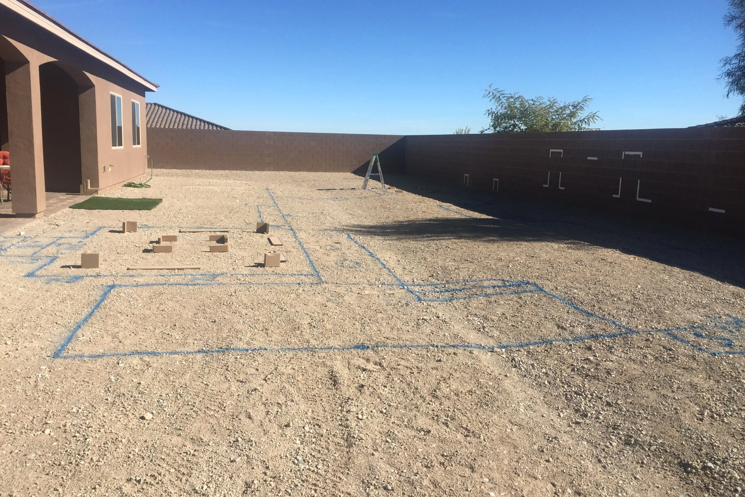 A backyard with gravel ground, blue chalk outlines for a basketball court, and a small construction area marked with blue chalk and wooden blocks. There is a brown house with a covered patio on the left and a tall, brown fence on the right. A ladder stands near the center of the yard, and a small patch of artificial grass is near the house. Clear blue sky overhead.