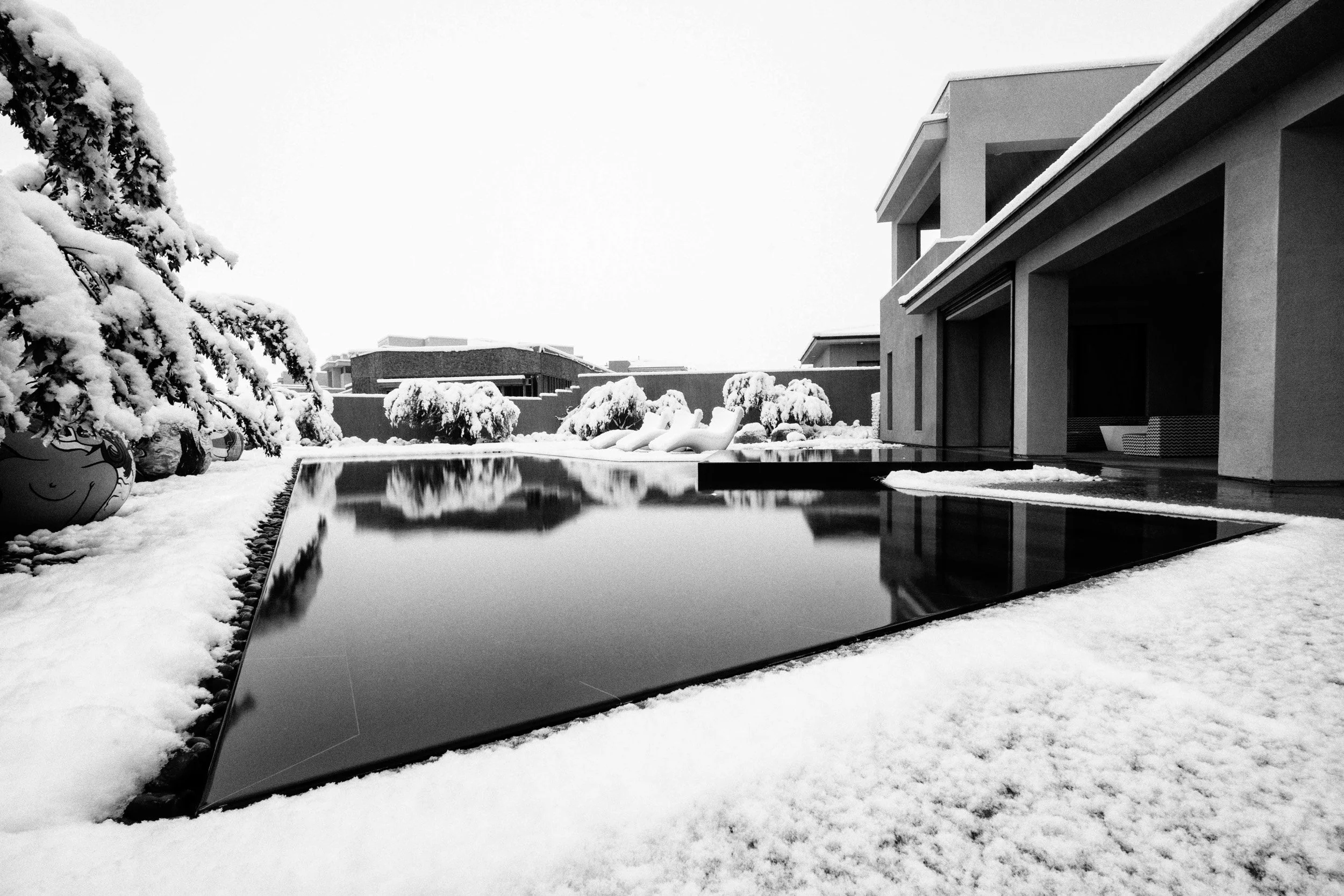 Modern house with a reflective pool in the snowy backyard, snow-covered trees, and outdoor lounge chairs.