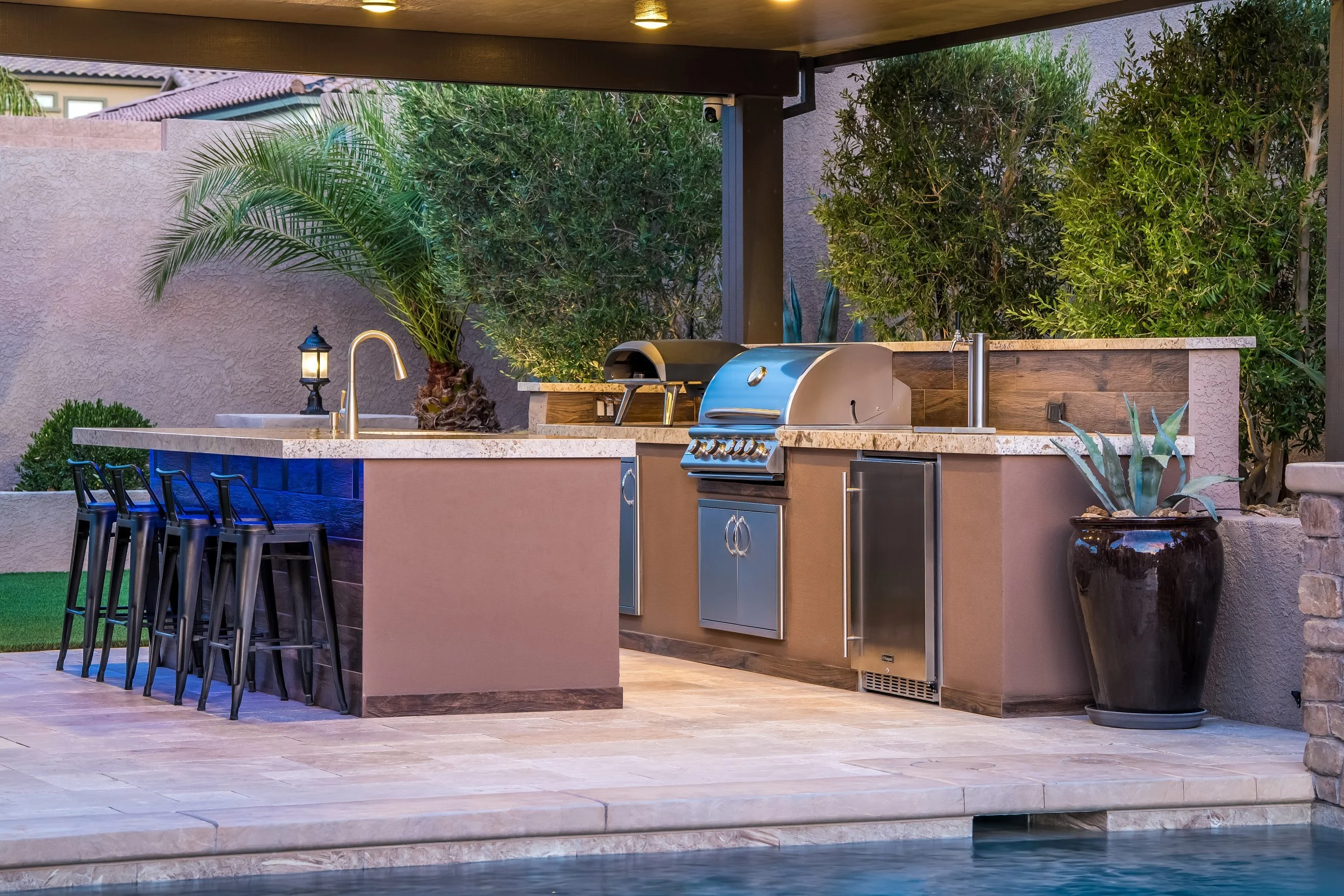 Outdoor kitchen with a granite countertop, stainless steel grill, beverages refrigerator, black bar stools, potted plant, palm tree, and lush greenery in a backyard setting
