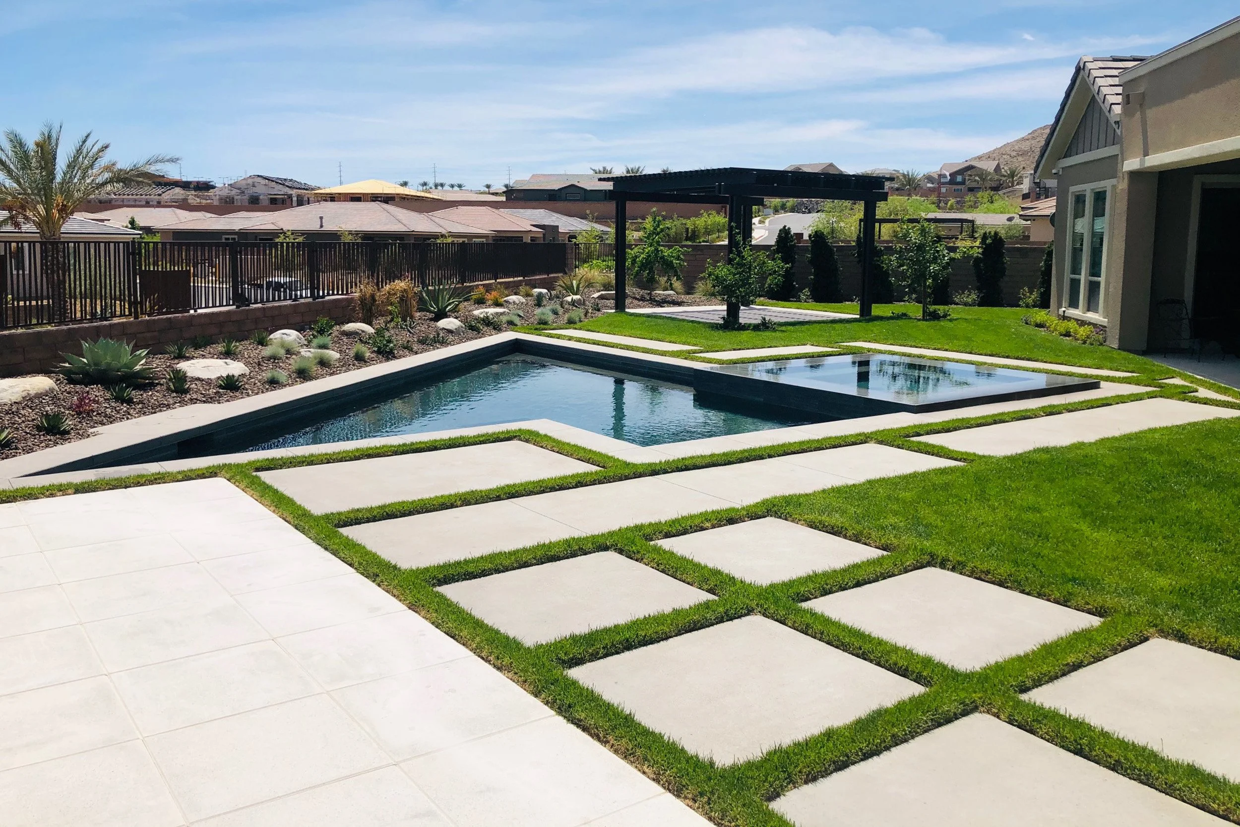 Backyard with a modern rectangular swimming pool, stone pathway, green grass, and landscaped plants under a clear blue sky.