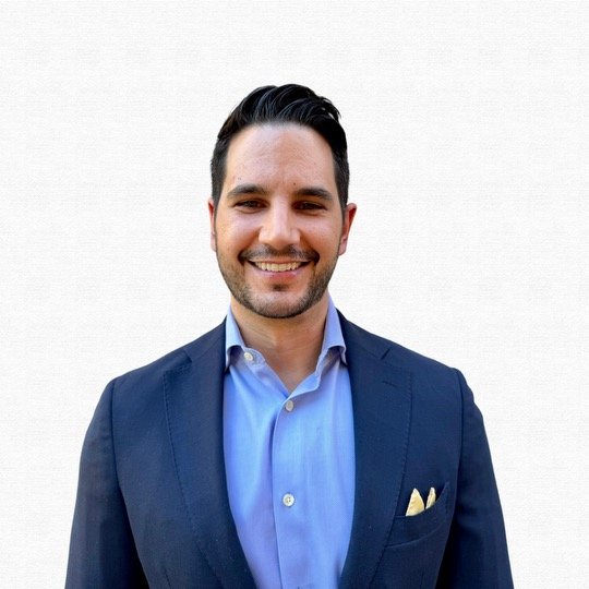 A smiling man with dark hair, wearing a blue suit and light blue shirt, standing against a plain light background.