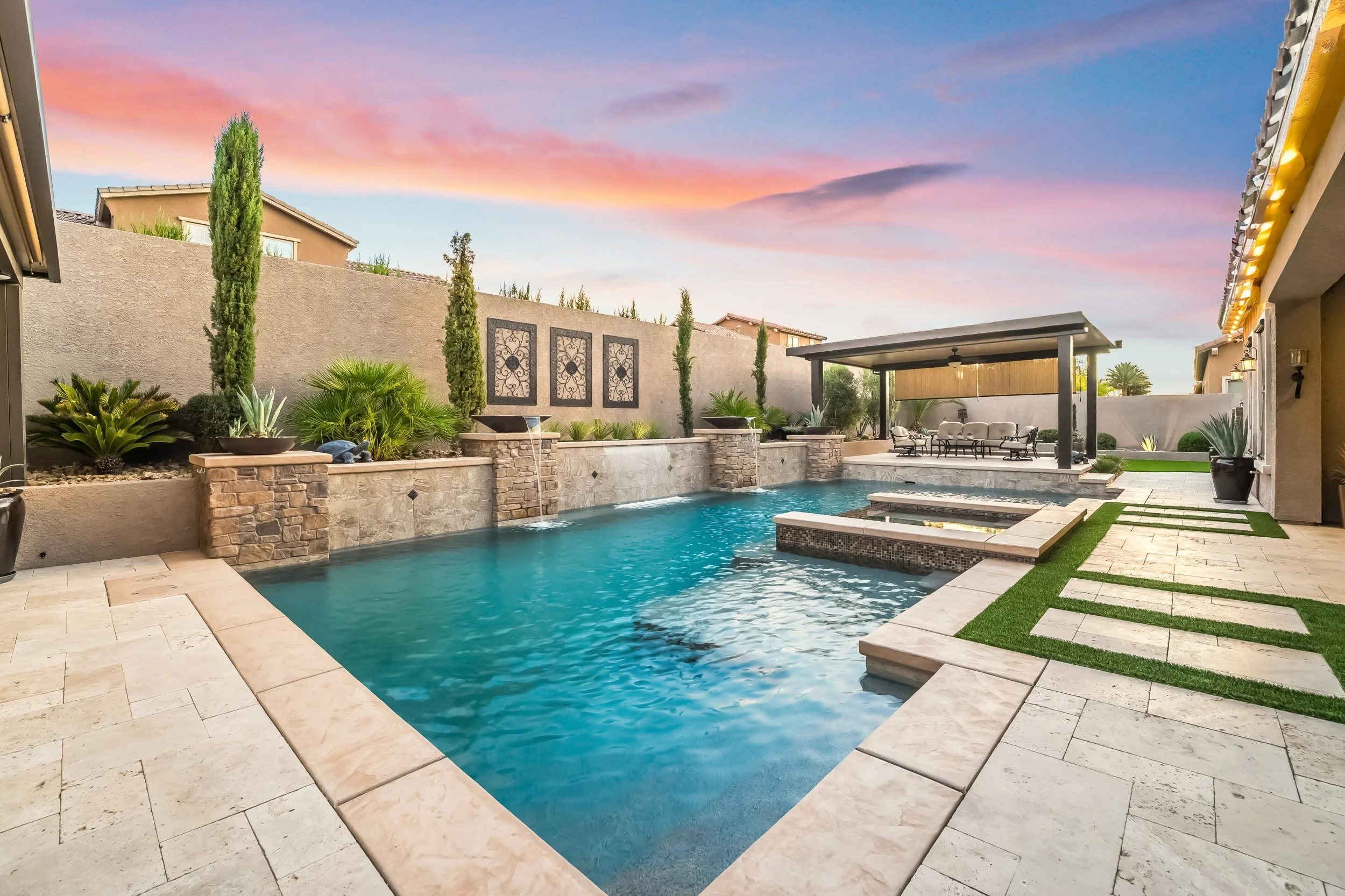 Backyard swimming pool with stone edging, waterfalls, covered seating area, lush greenery, and sunset sky.
