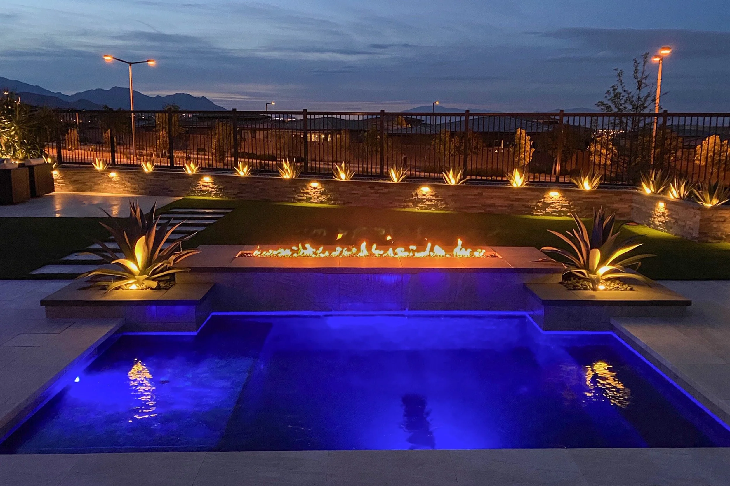 Nighttime backyard with a pool illuminated by blue lights, a fire feature in the center, and landscaped plants with lights along the fence. Mountains are visible in the distance.
