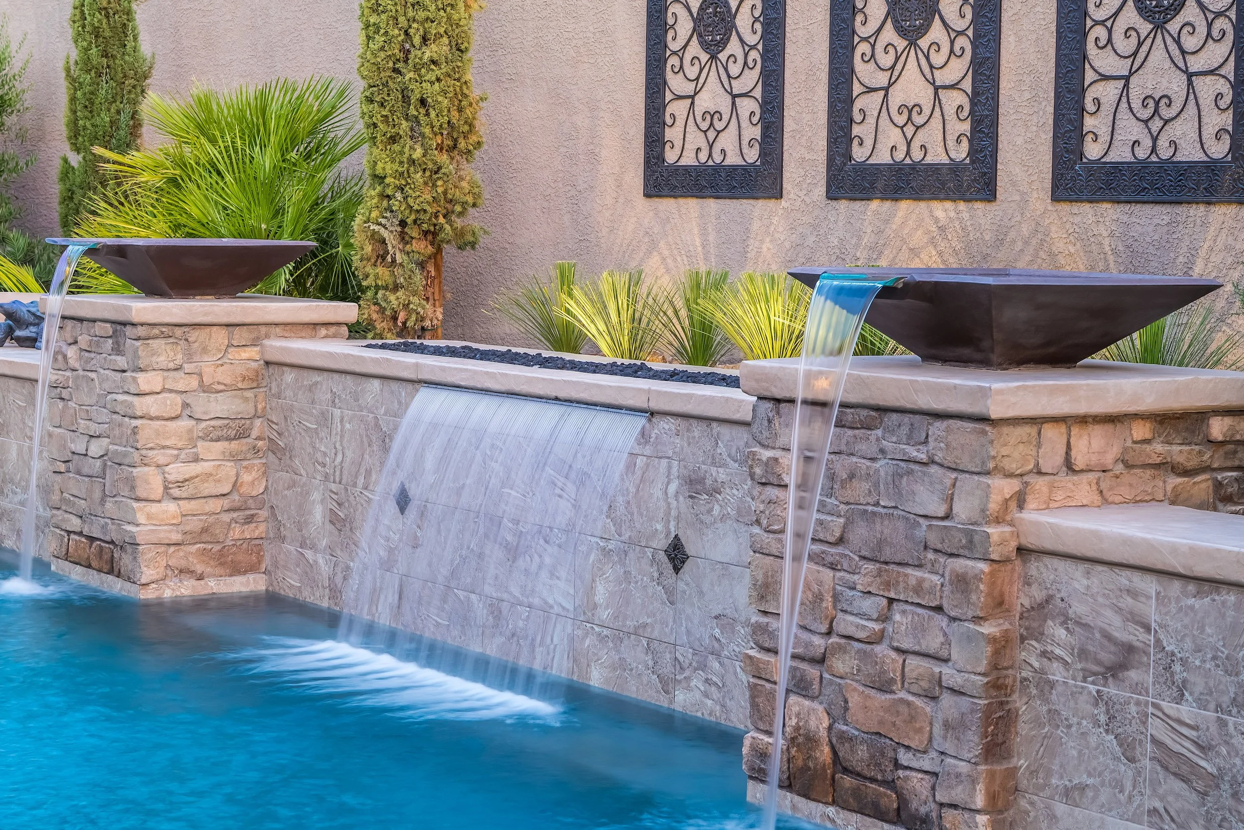 A backyard fountain with two modern water bowls on stone pedestals and a waterfall from a tiled wall, surrounded by lush green plants and a textured wall with decorative iron window grates.
