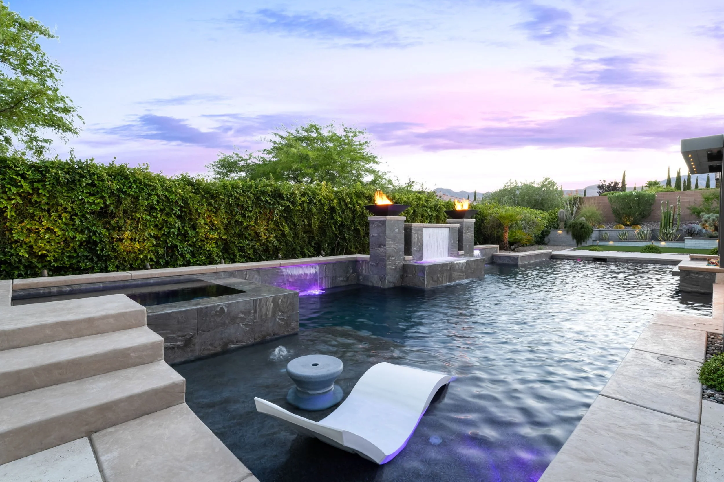 Luxury backyard swimming pool with built-in seating, waterfall, stone fire pits, surrounded by lush green bushes and decorative plants, during sunset.