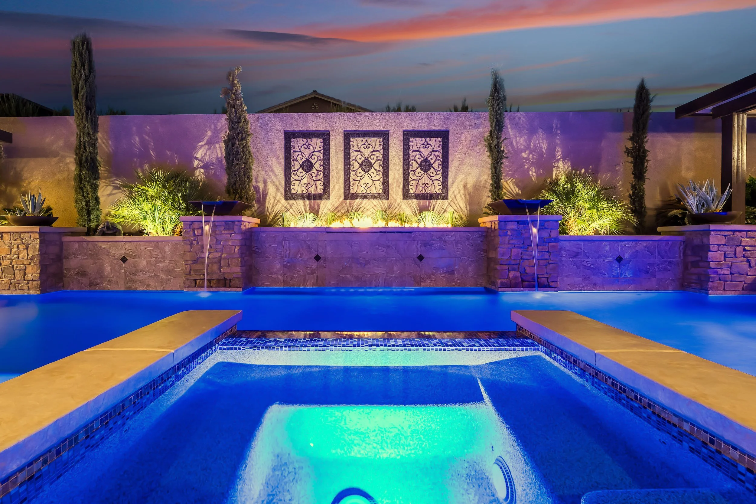 An illuminated backyard swimming pool at twilight with a decorative stone wall featuring three ornate metal panels, surrounded by tall cypress trees and lush plants, creating a serene ambiance.