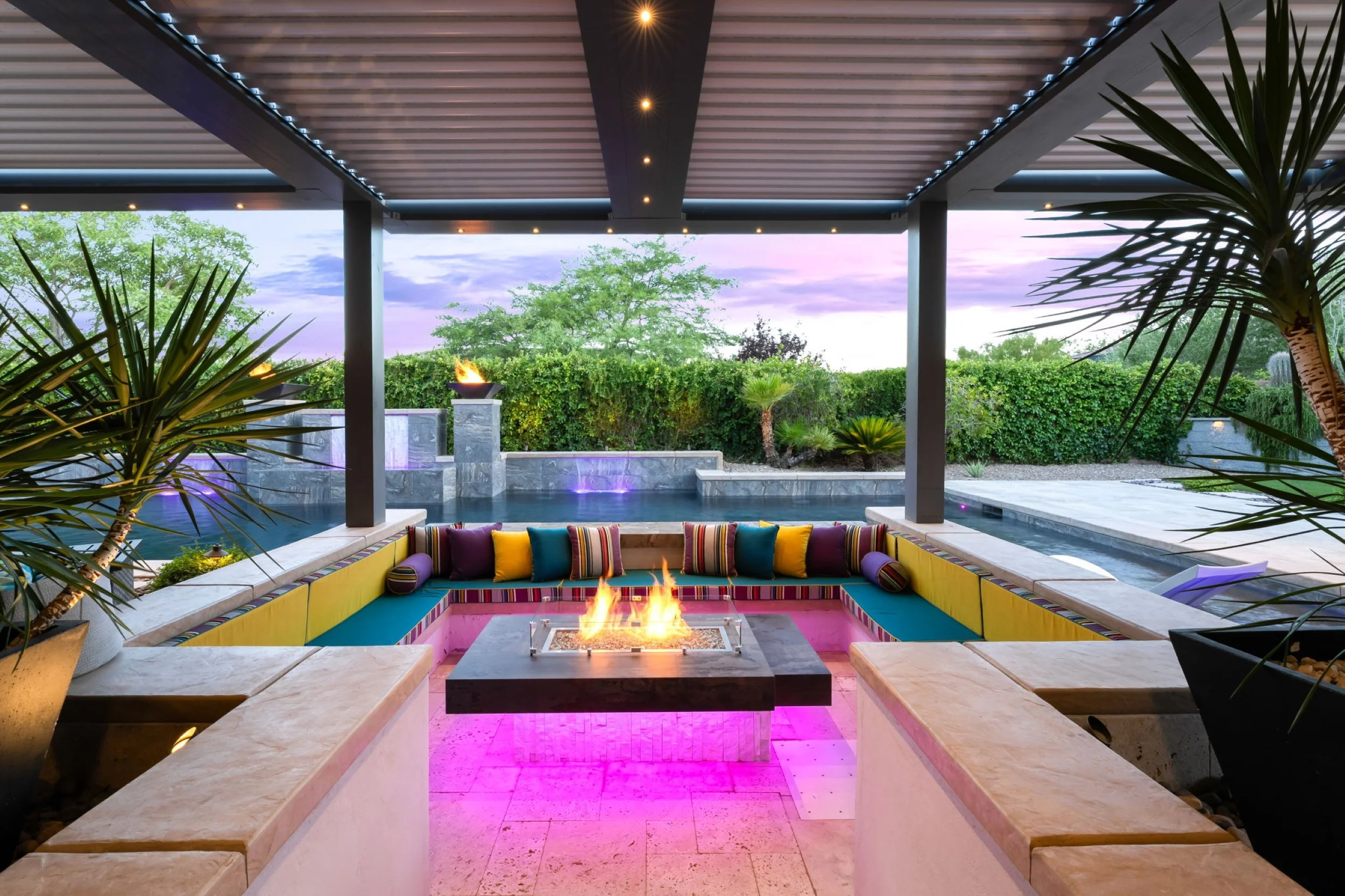 Covered outdoor patio with a seating area with colorful cushions, a central fire pit, a pool with a fire feature, tropical plants, and a sunset sky in the background.