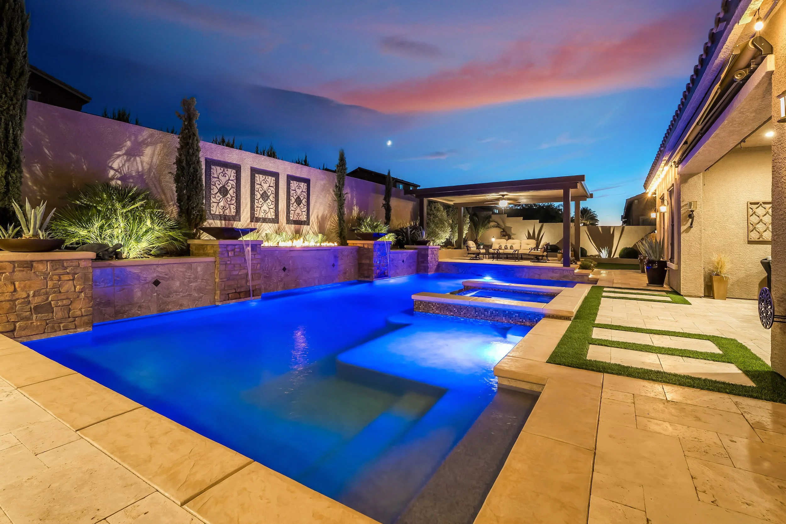 A backyard with a swimming pool illuminated at night, surrounded by stone and tile decks, with plants and outdoor seating areas, under a twilight sky.