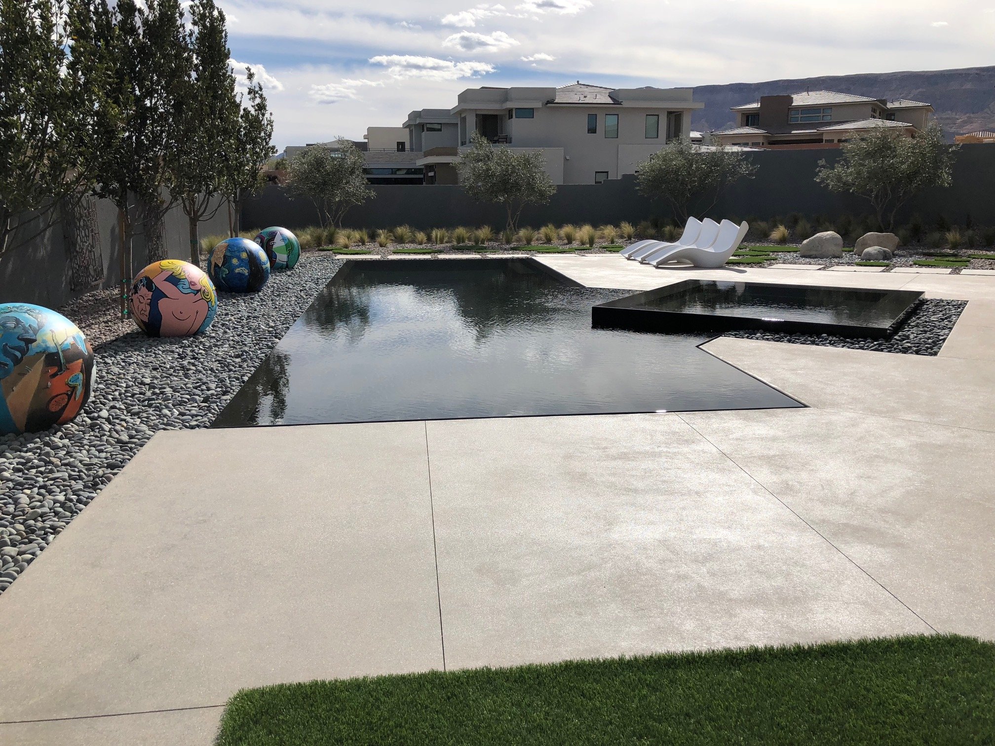 Modern backyard pool area with white pool chairs, artistic painted spherical sculptures, and nearby trees with a mountain in the background.