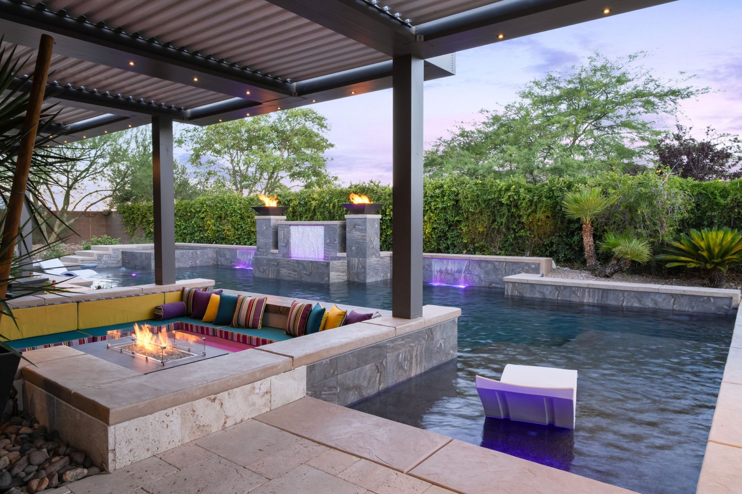 Outdoor pool area with a fire pit, colorful seating area, water feature with flame bowls, and surrounding greenery in the evening