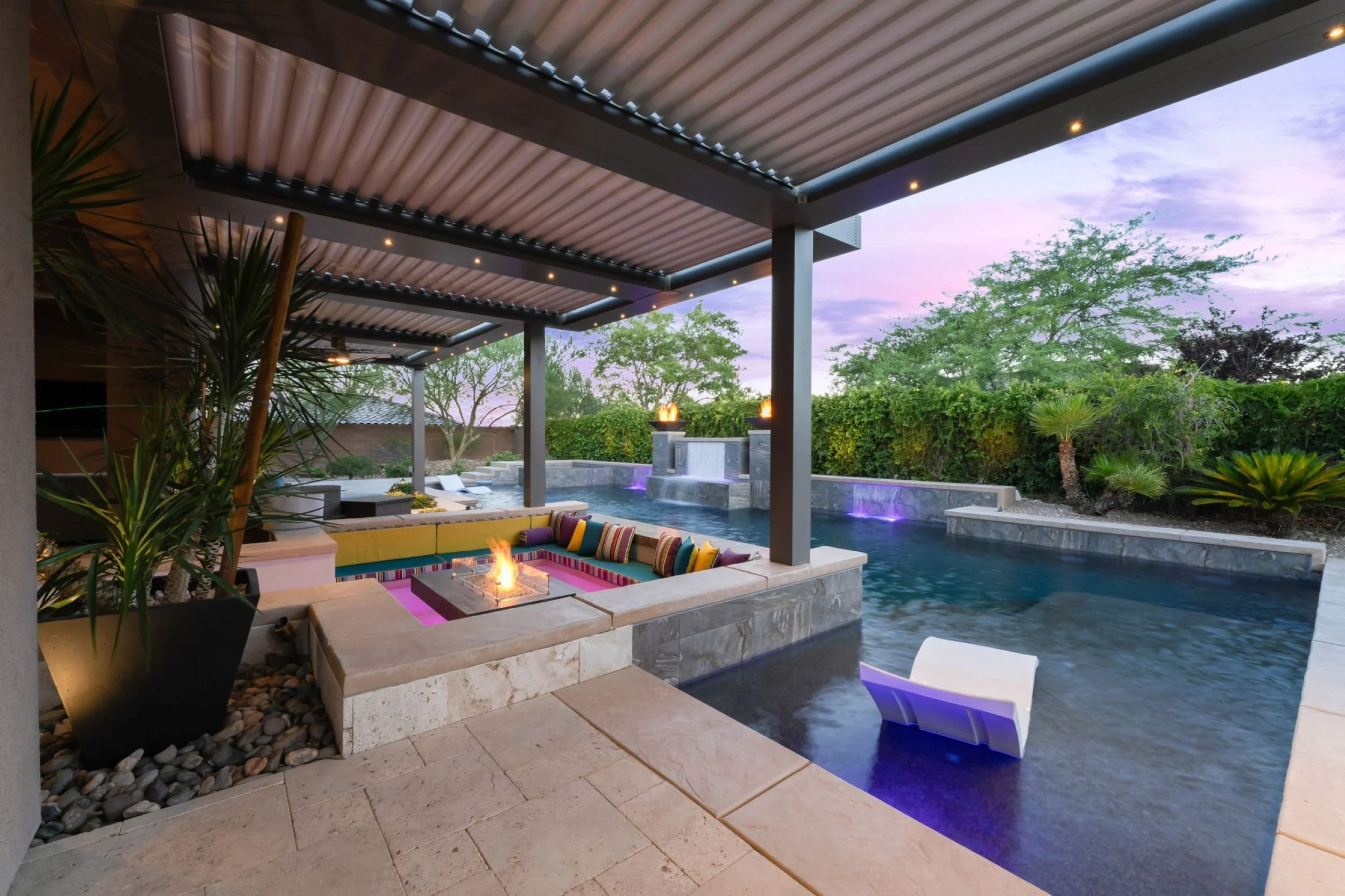 Luxury backyard with swimming pool, outdoor fire pit with seating, waterfalls, colorful lighting, and lush greenery at dusk.