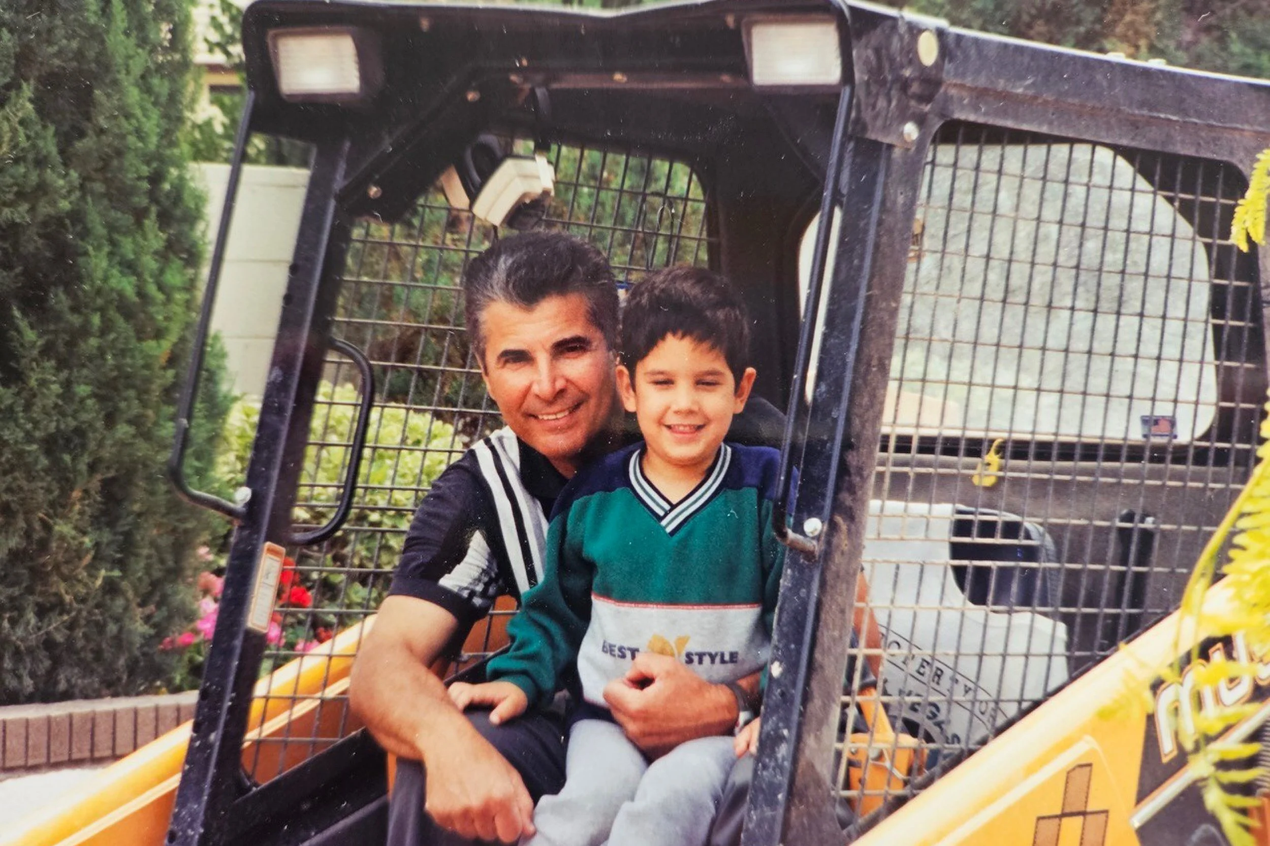 A man and a young boy sitting inside a yellow utility vehicle or construction vehicle, smiling at the camera. The background includes bushes and trees.