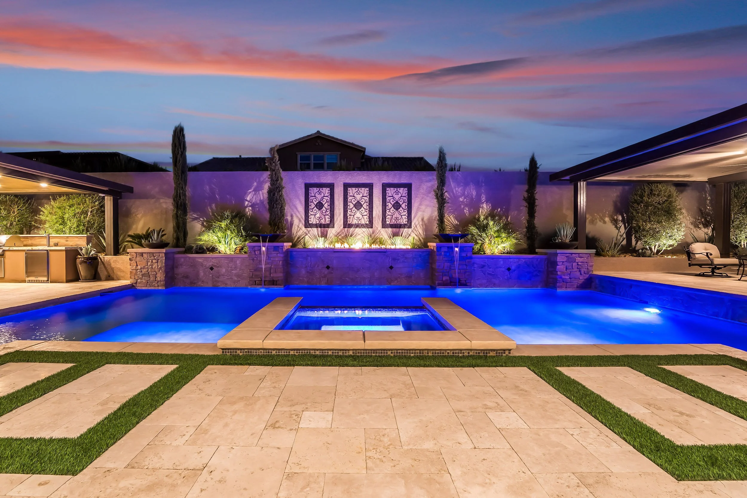 A luxurious backyard swimming pool area illuminated at night with blue lighting and decorative landscaping, including trees and potted plants, surrounded by a patio and seating area.